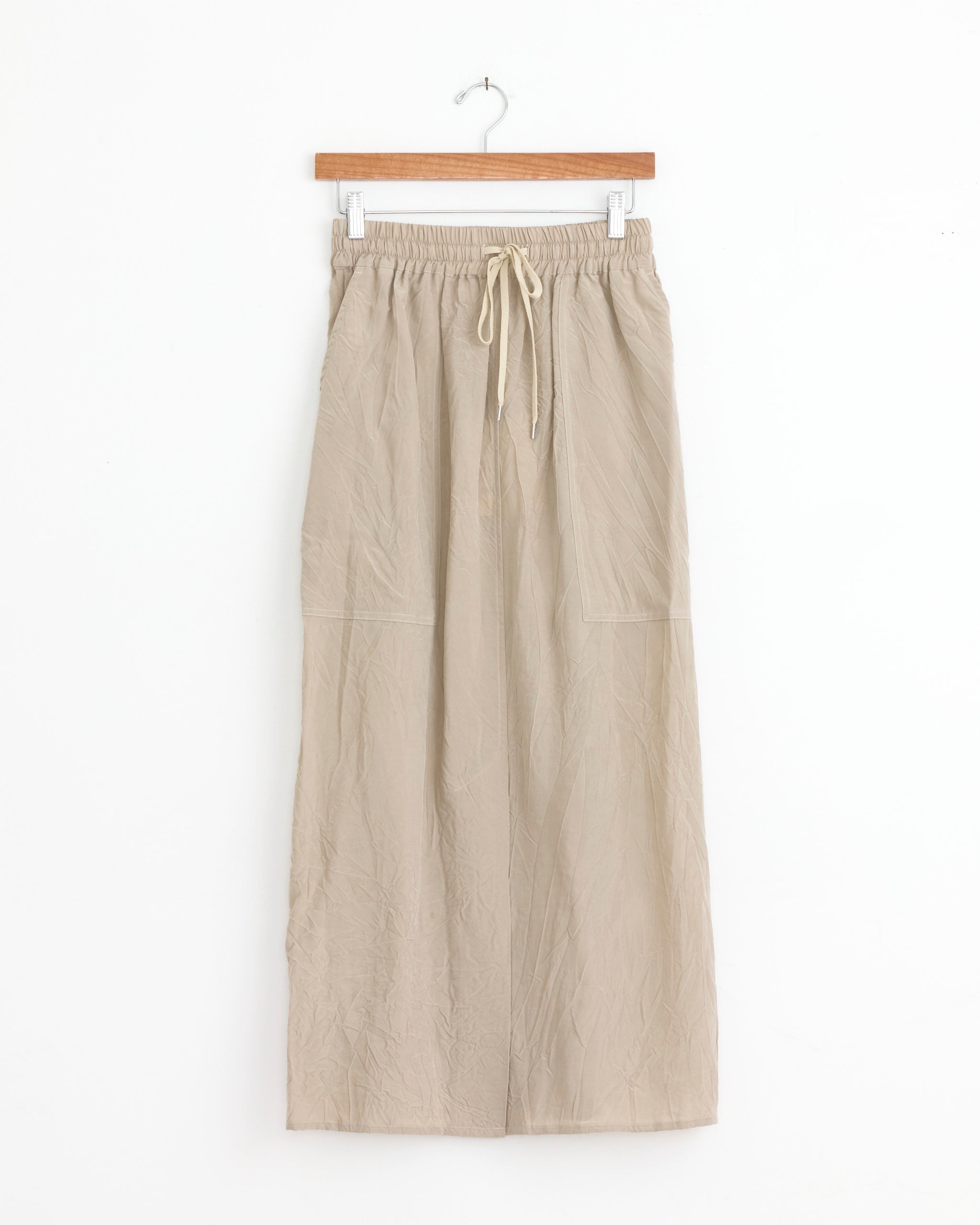 The Oates Skirt in Sable by Studio Nicholson features a beige wide-leg, ankle-length design with an elasticated waistband, drawstring tie, and two large Italian patch pockets. Displayed on a wooden hanger against a white background with subtle workwear touches.