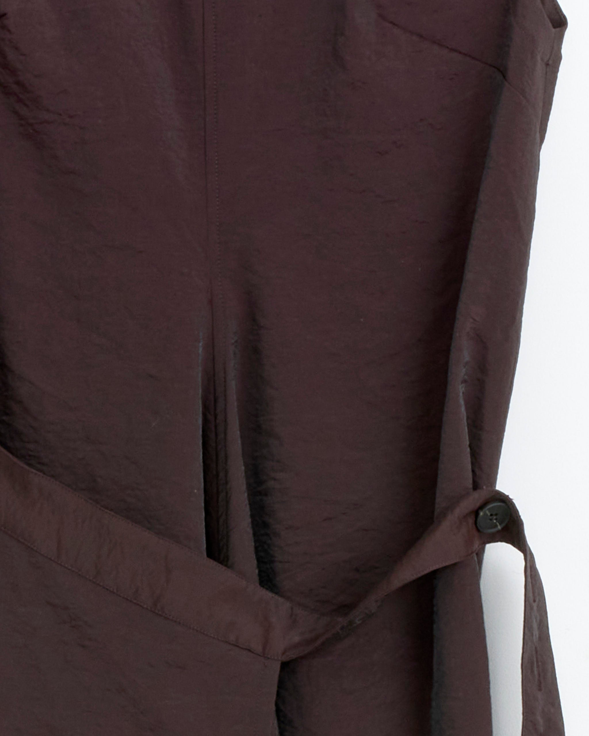 Close-up of Studio Nicholson’s Verve Dress in Cocoa—a dark brown, textured wool-nylon blend—showing a visible seam, a buttoned strap, and part of a pocket against a white background.