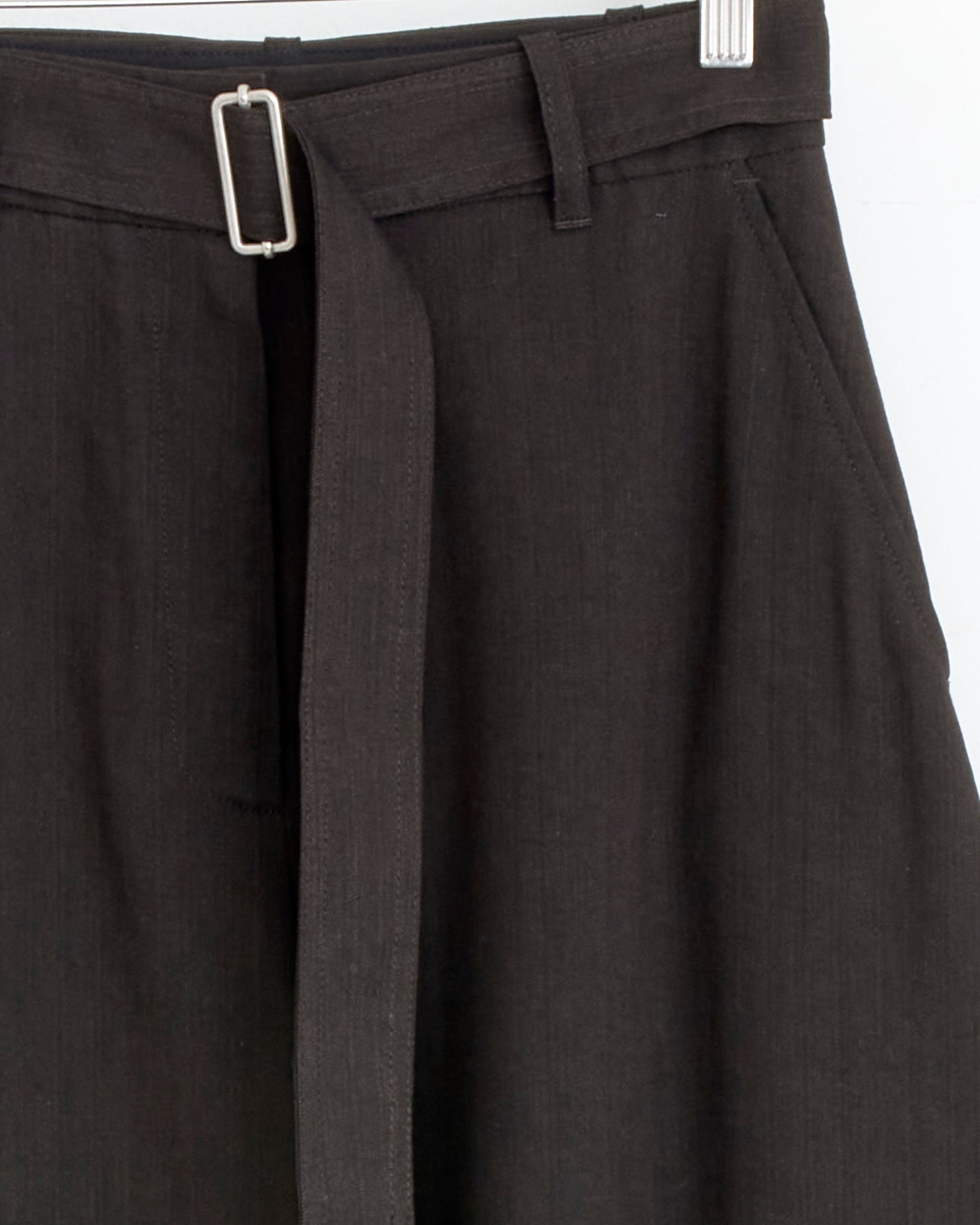 Close-up of the Studio Nicholson Chatto Pant in Espresso, featuring herringbone fabric with a matching belt and silver rectangular buckle, displayed on a hanger against a plain white background.