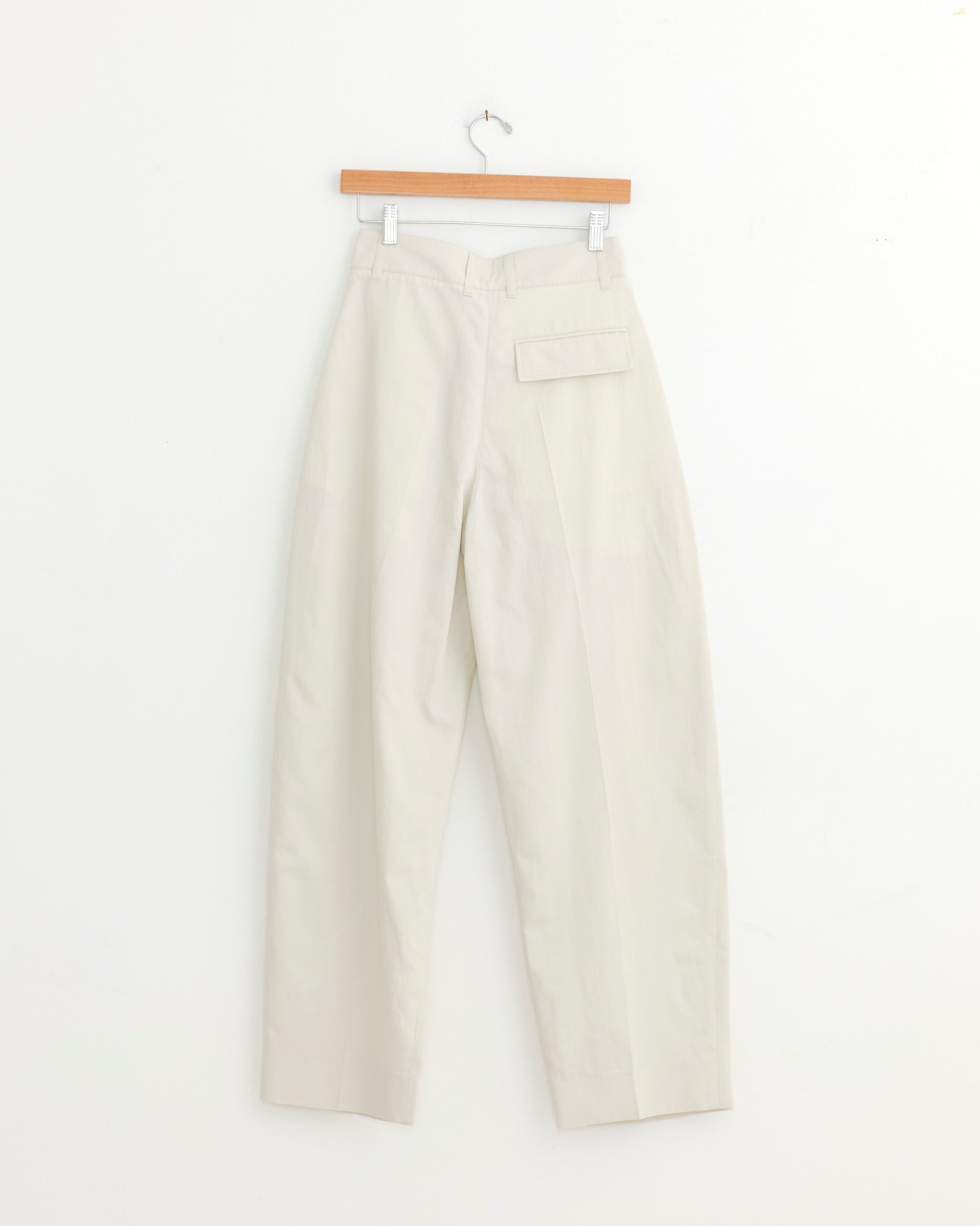 The Studio Nicholson Acuna Pant in Bone, a high-rise wide-leg style made from Italian cotton-viscose twill, hangs on a wooden hanger. It features a single flap pocket on the back right side, set against a plain white background.