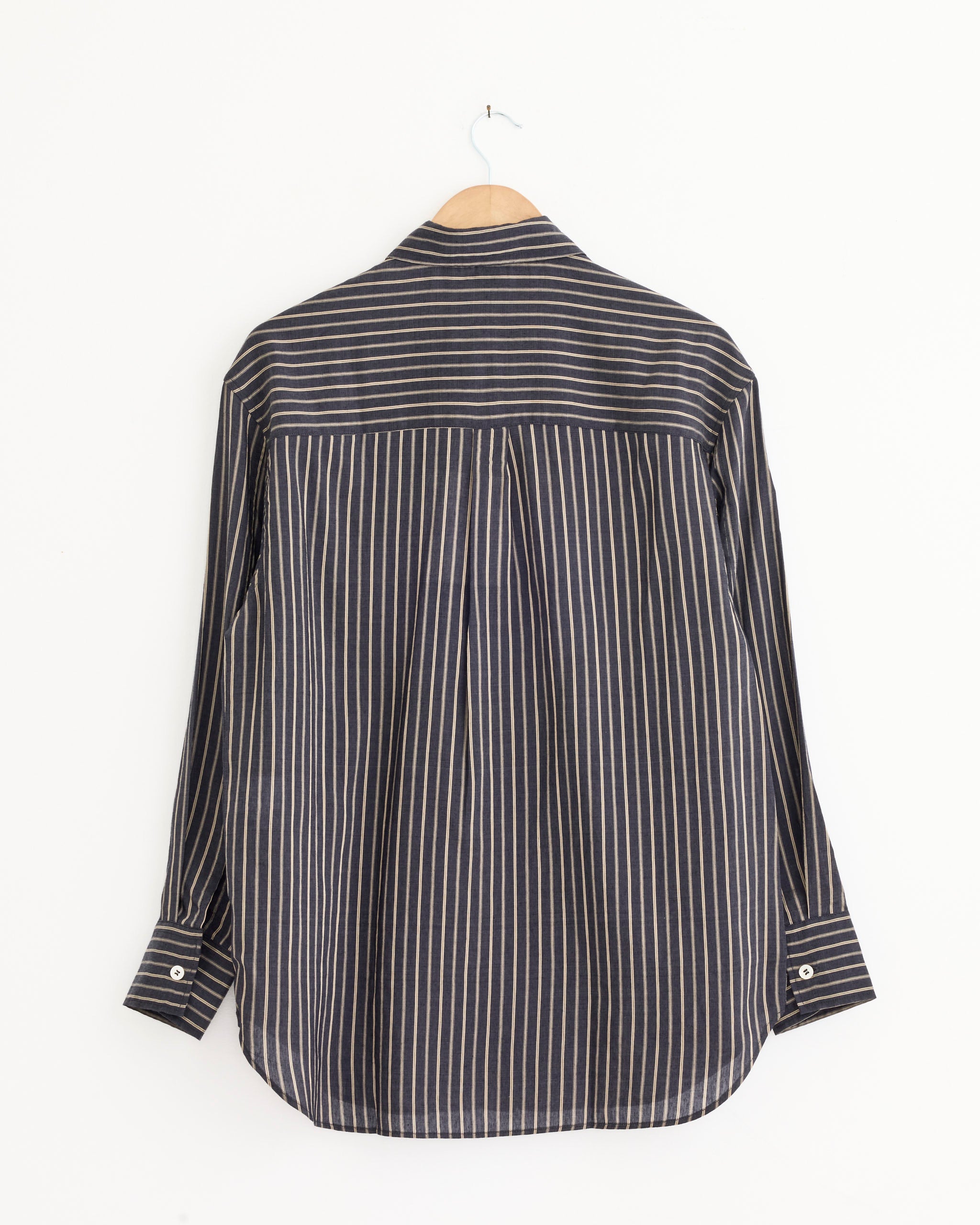 The Studio Nicholson Rouen Shirt in Indigo, a contemporary long-sleeved shirt with white vertical stripes, is displayed from the back on a wooden hanger against a plain white background.