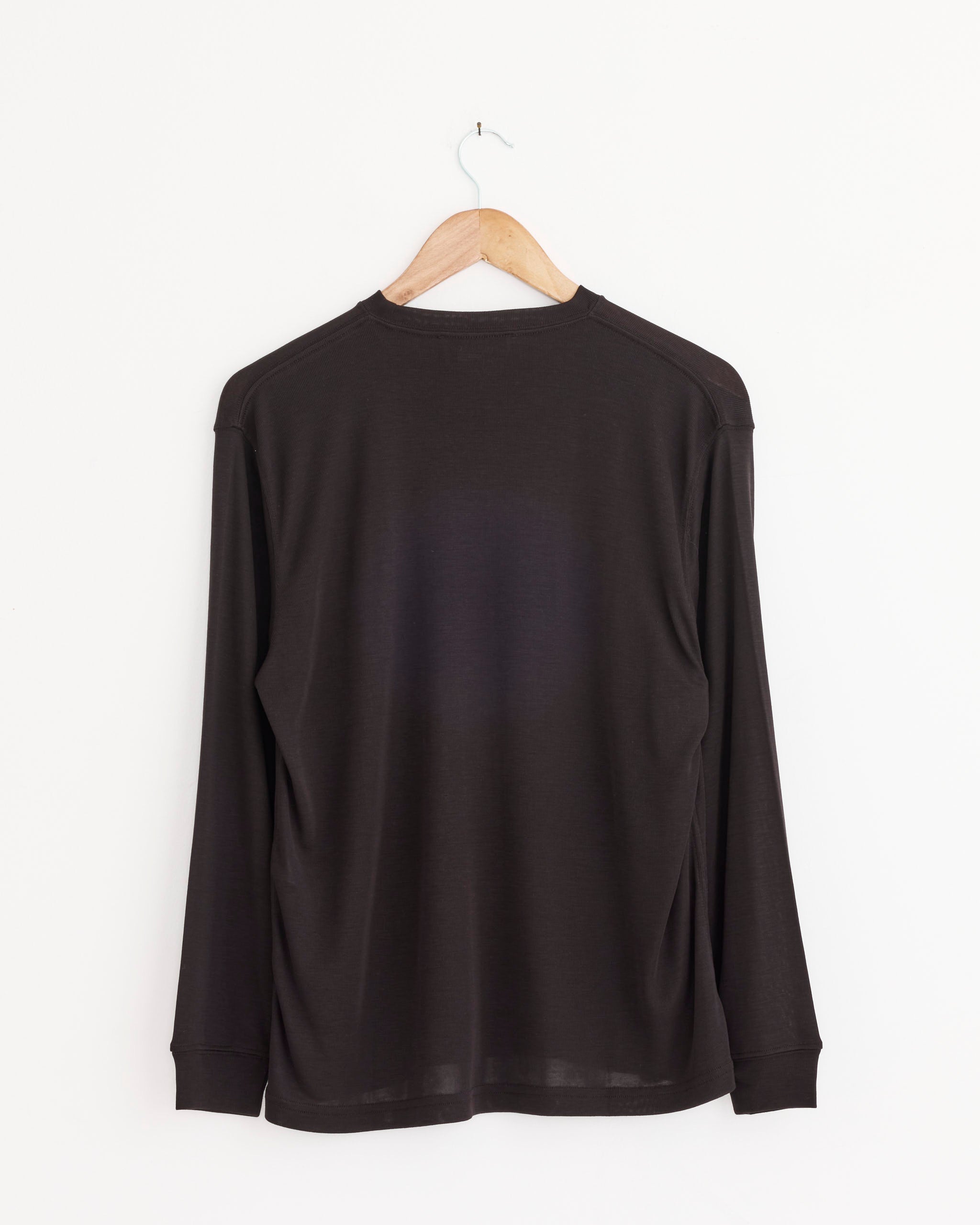 The Simmons T-Shirt in Black Grape by Studio Nicholson, featuring long sleeves and an oversized fit, hangs on a wooden hanger against a plain white wall, displayed from the back.