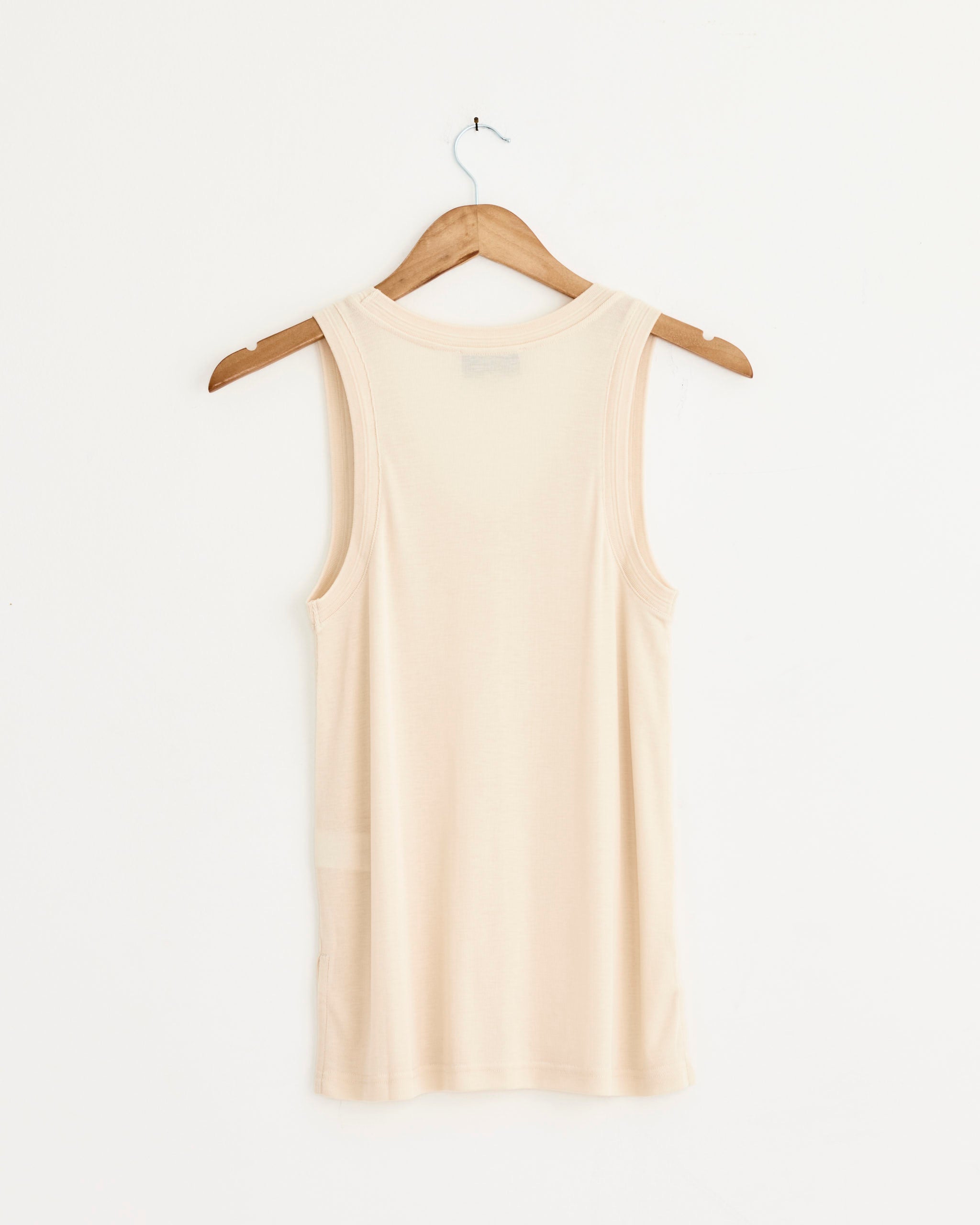 The Studio Nicholson Votan Vest in Canvas, a sleeveless beige lyocell tank with a relaxed fit, is displayed on a wooden hanger against a plain white wall and shown from the back.