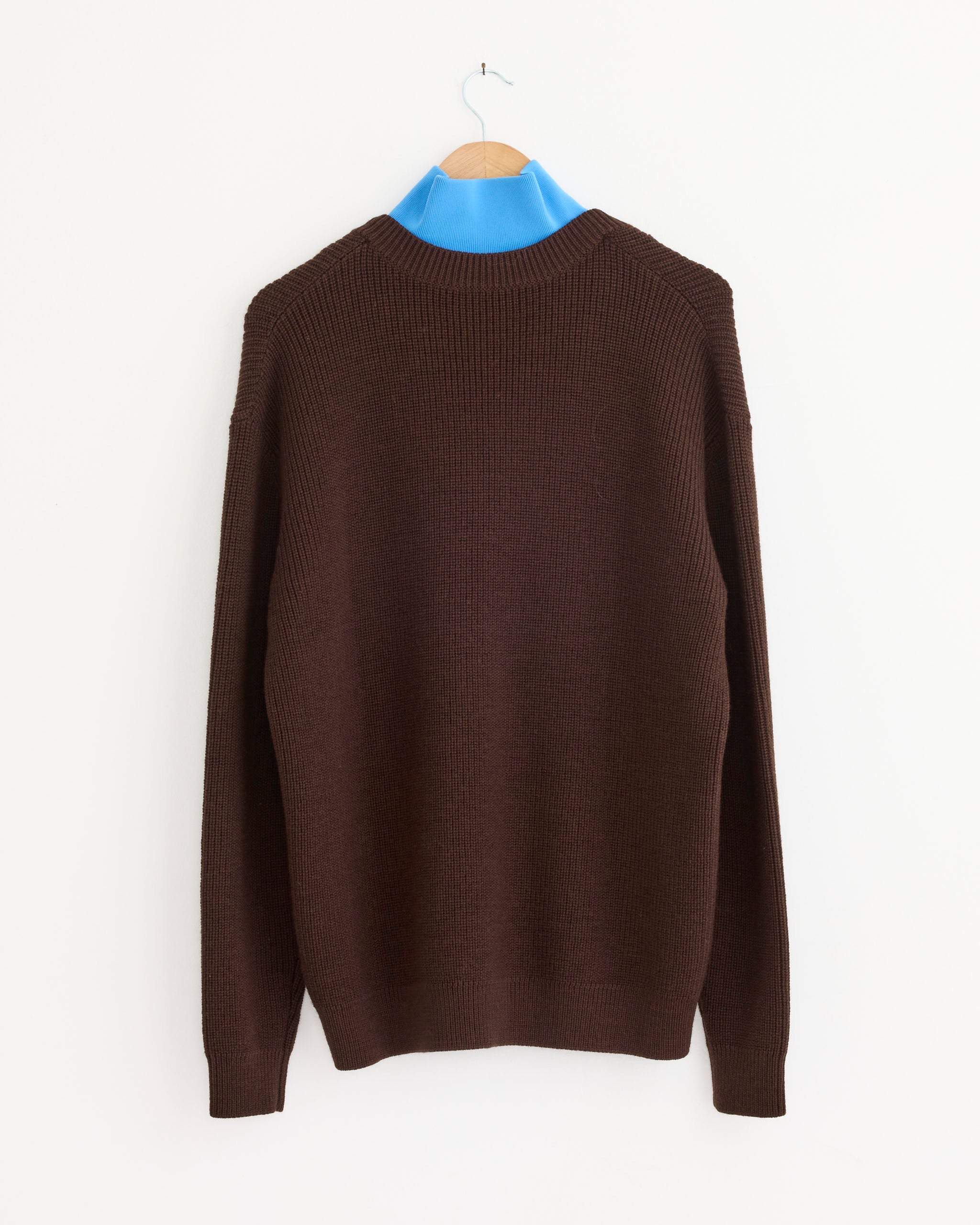 The V Neck Cardigan in Brown by Tibi hangs on a wooden hanger against a white wall, styled over a blue turtleneck shirt for extra warmth and sophistication.