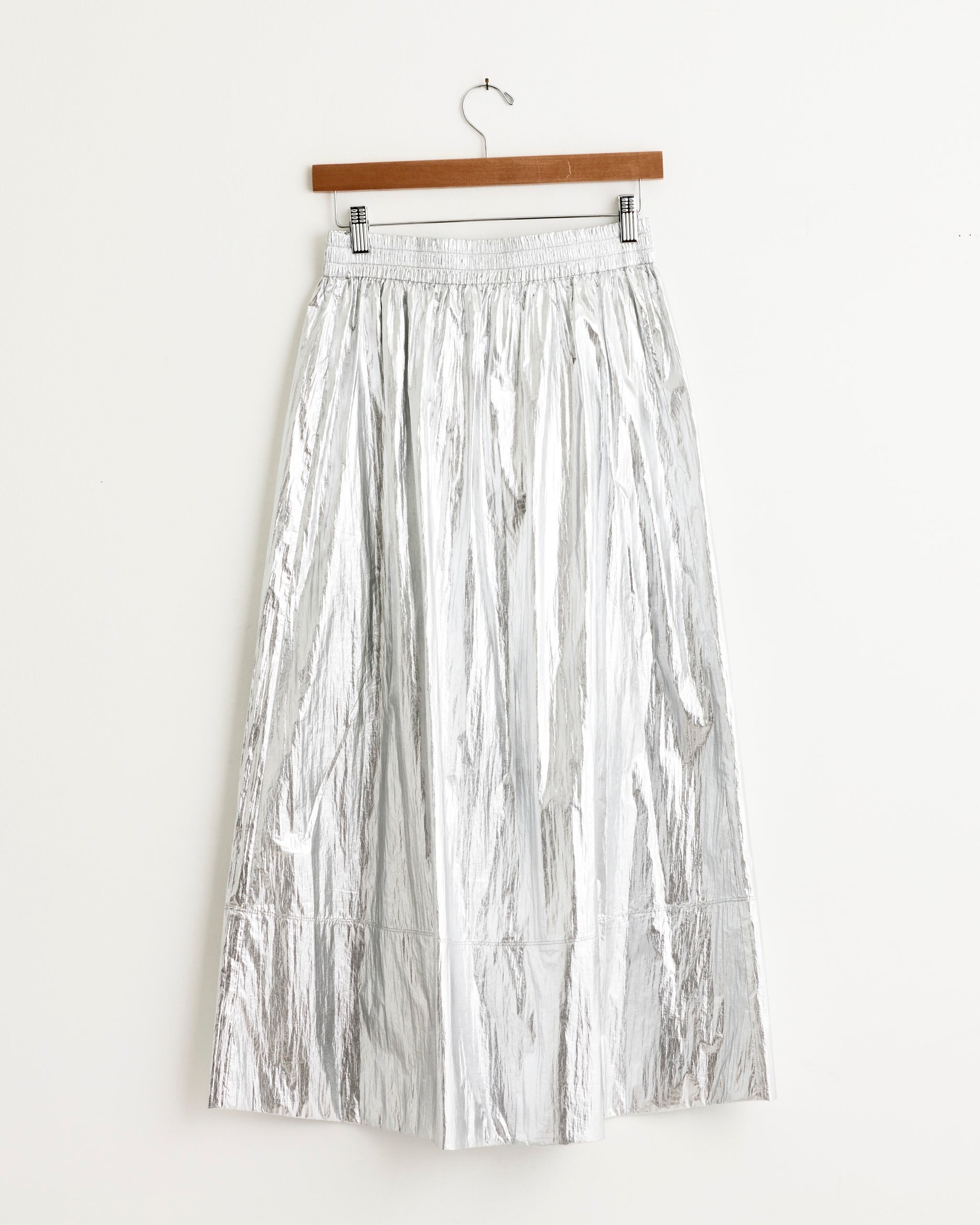 The Tibi Metallic Nylon Pull On Skirt in Silver, with a high-shine finish, hangs on a wooden hanger against a plain white wall.
