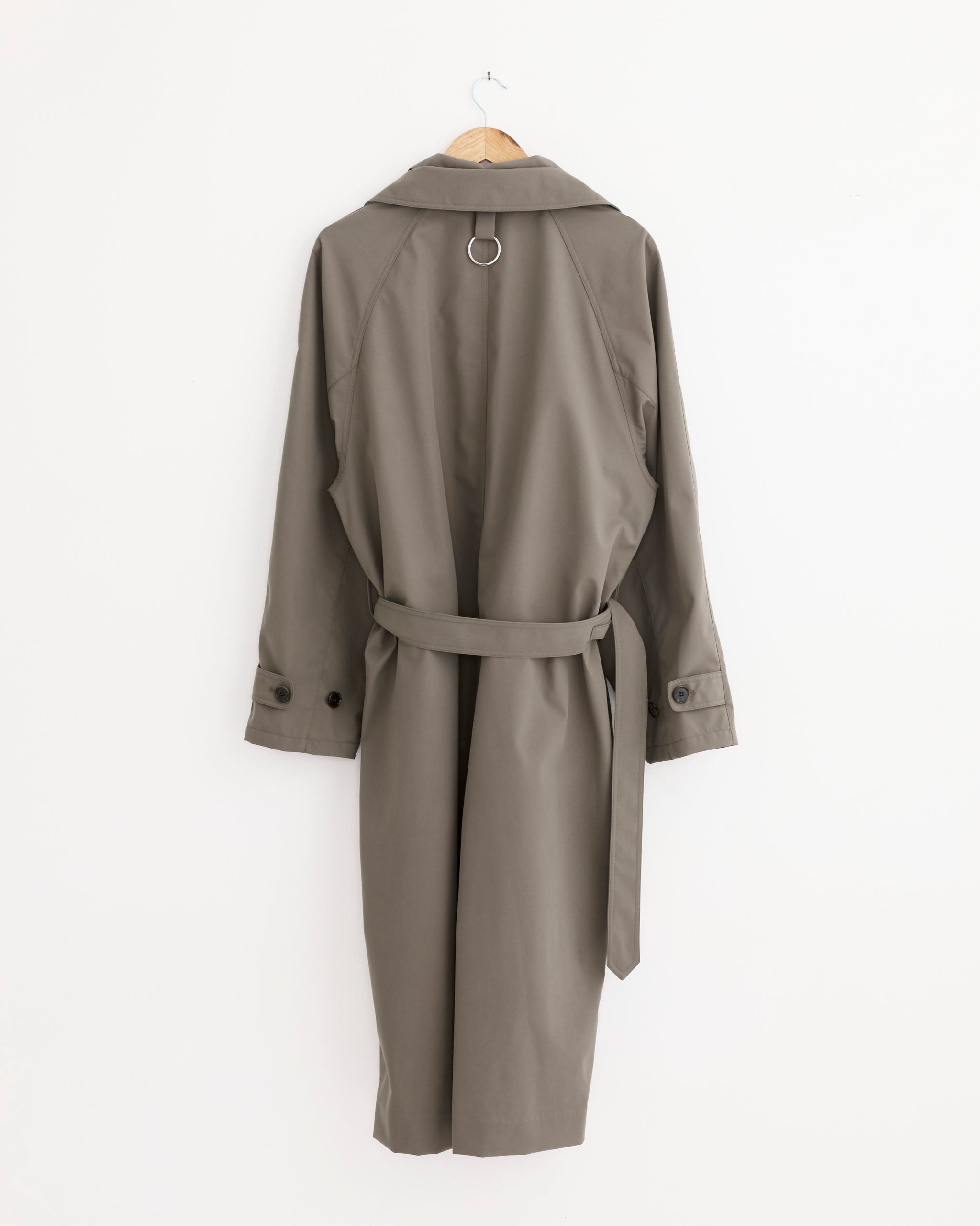 The Tibi Double Collar Frank Trench Coat in Dark Hazelnut, shown on a wooden hanger against a white wall, features a detachable waist belt, buttoned cuffs, and a metal ring detail on the back yoke.