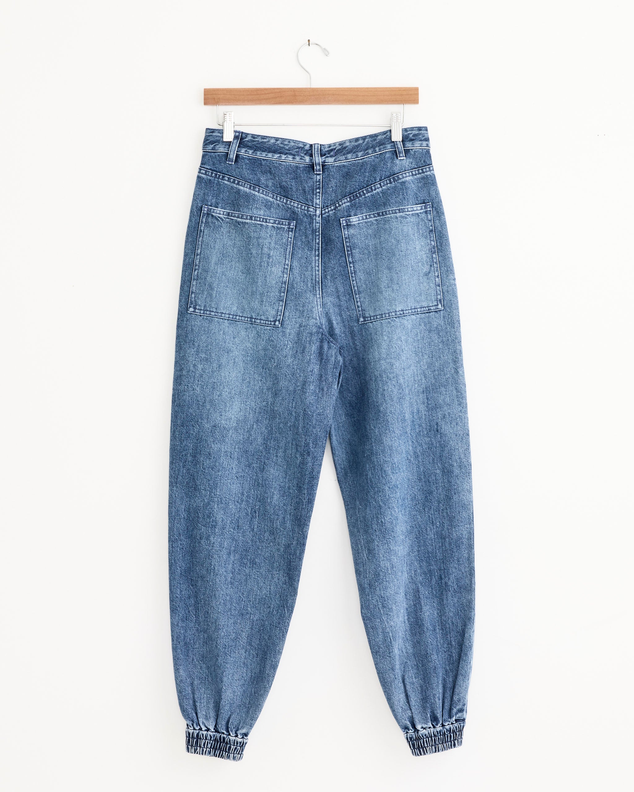 The Tibi Zippered Denim Jogger in Classic Blue, featuring a high waist, elasticated cuffs, and large back pockets, hangs on a wooden hanger against a plain white background.