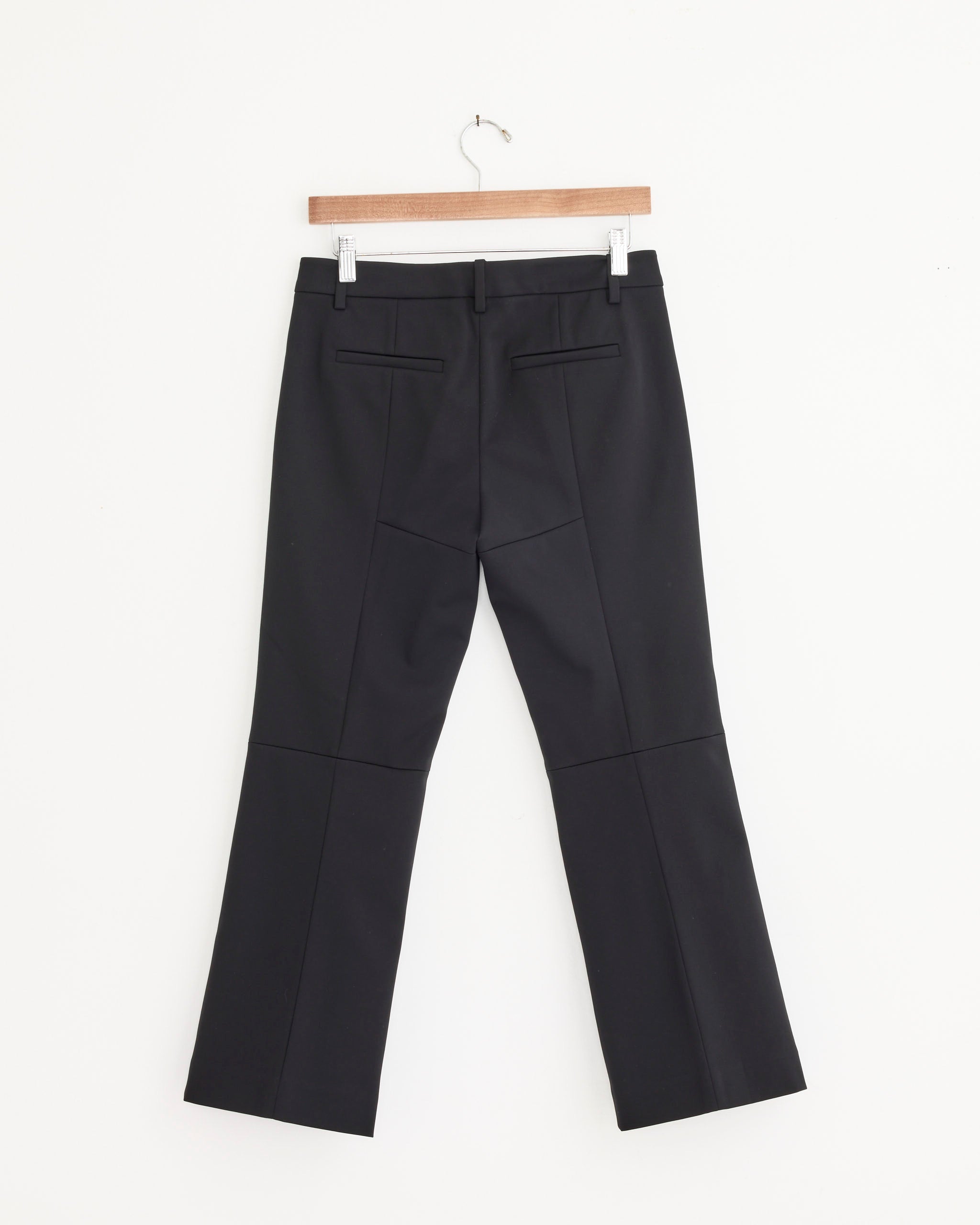 The Tibi Compact Tech Knit Bootcut Pant in Black hangs on a wooden hanger against a white wall, featuring a cropped silhouette and back welt pockets.