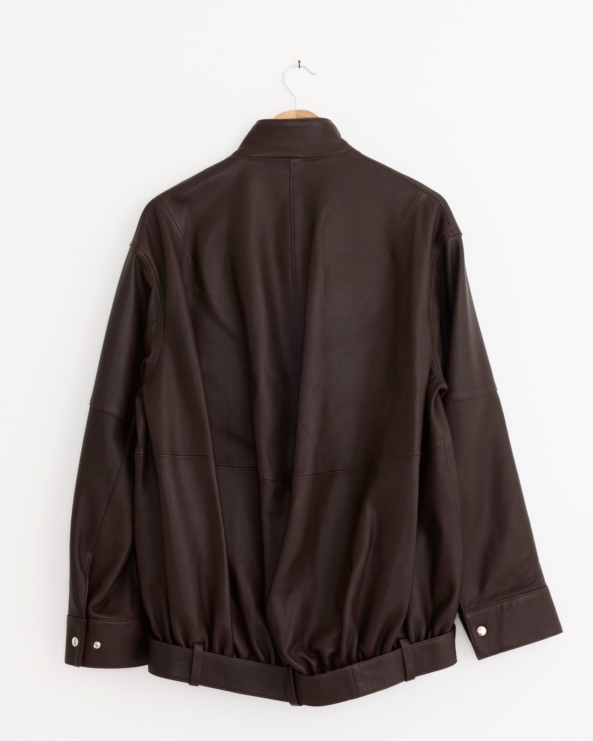 The Tibi Leather Moto Bomber Jacket in Brown—a statement lamb leather piece—hangs on a white wall, shown from the back. It features long sleeves with buttoned cuffs, a belted waist, and a simple collar.