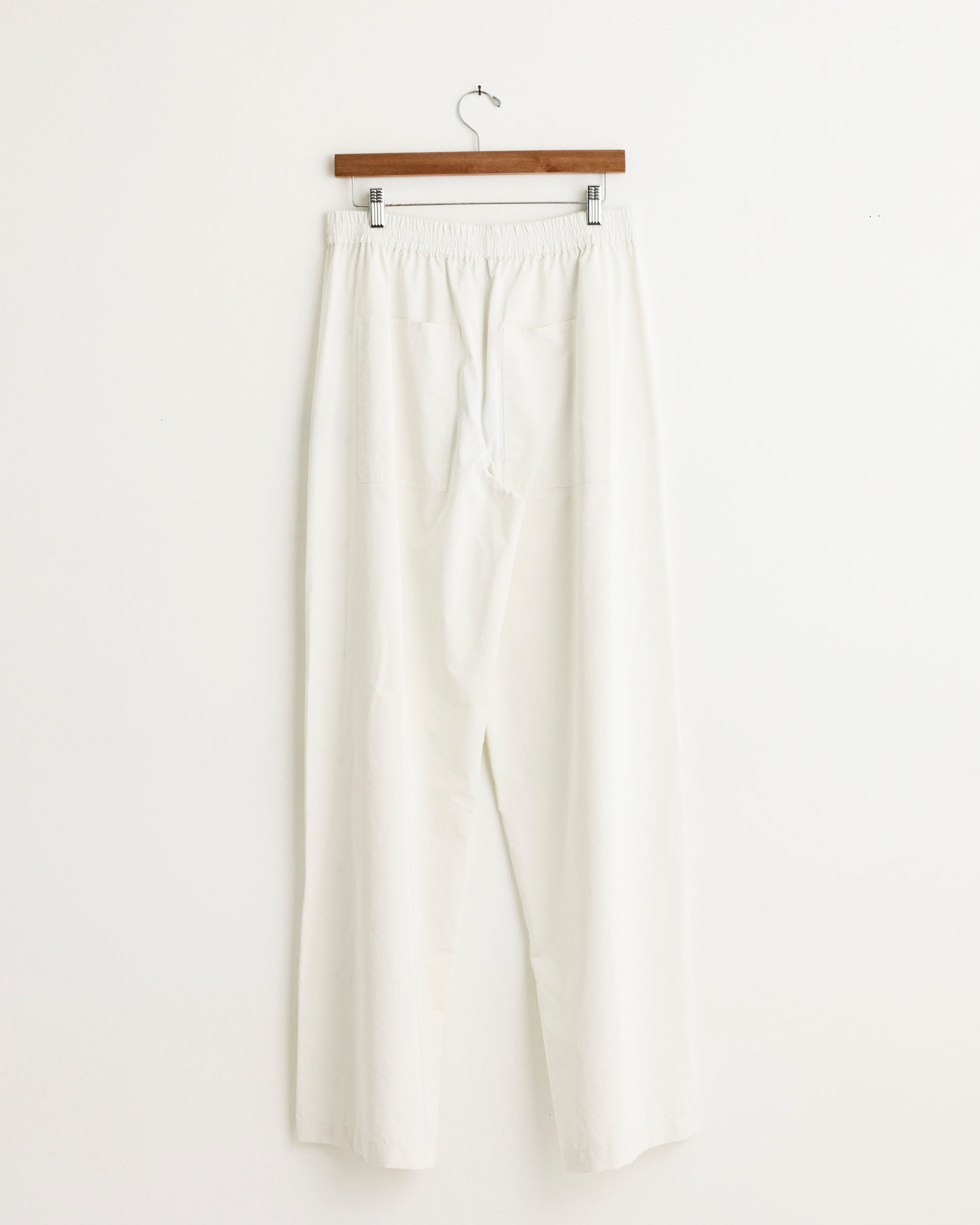 The Tibi Nylon Snap Pull On Jogger in White features a loose fit, wrinkle-resistant fabric, and adjustable waistband with drawstring, displayed hanging on a wooden hanger against a plain white wall.