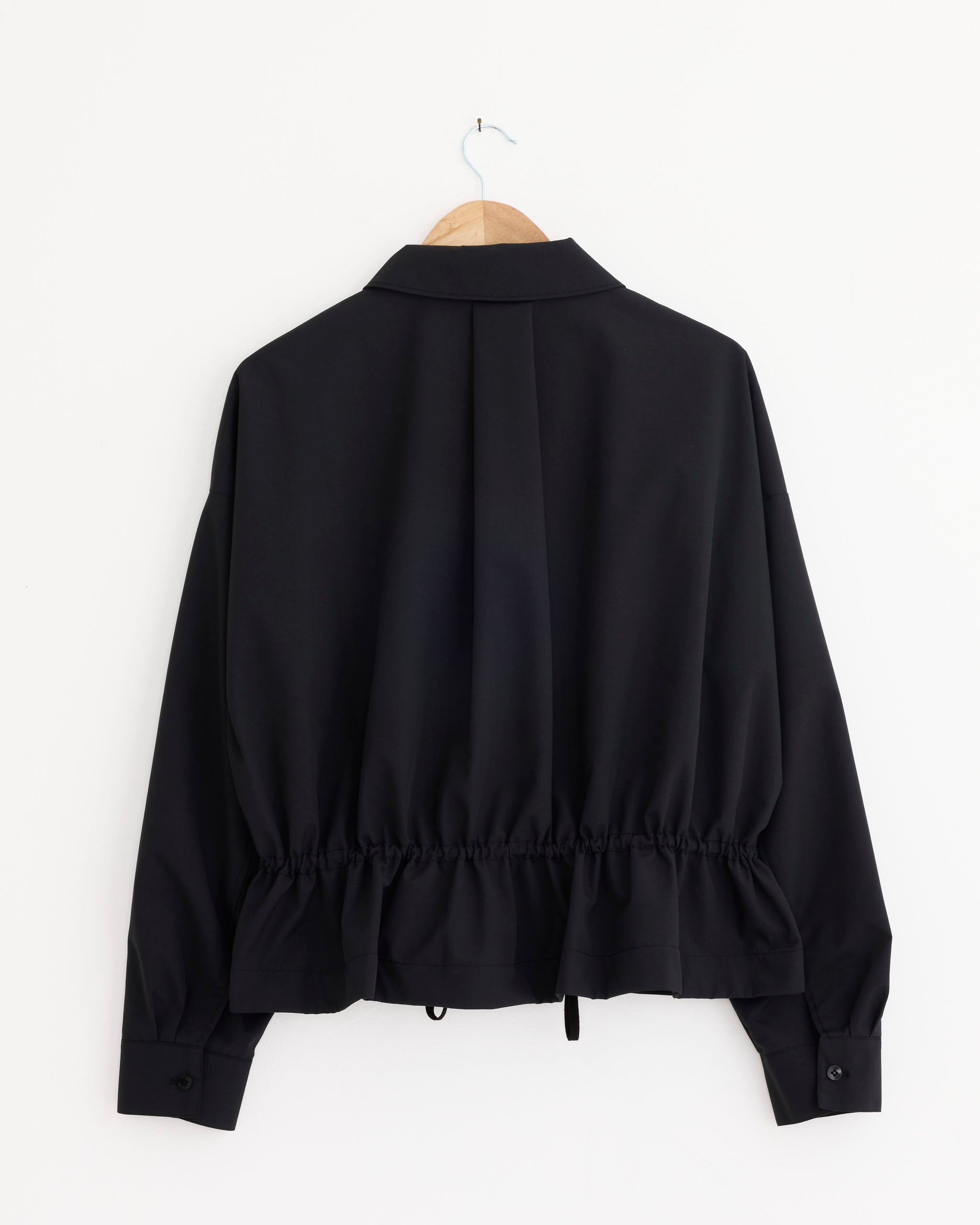 The Tibi Tech Suiting Anorak Bomber Jacket in Black, featuring relaxed tailoring, a gathered waist, adjustable drawstring, and buttoned cuffs, hangs on a wooden hanger against a white background.