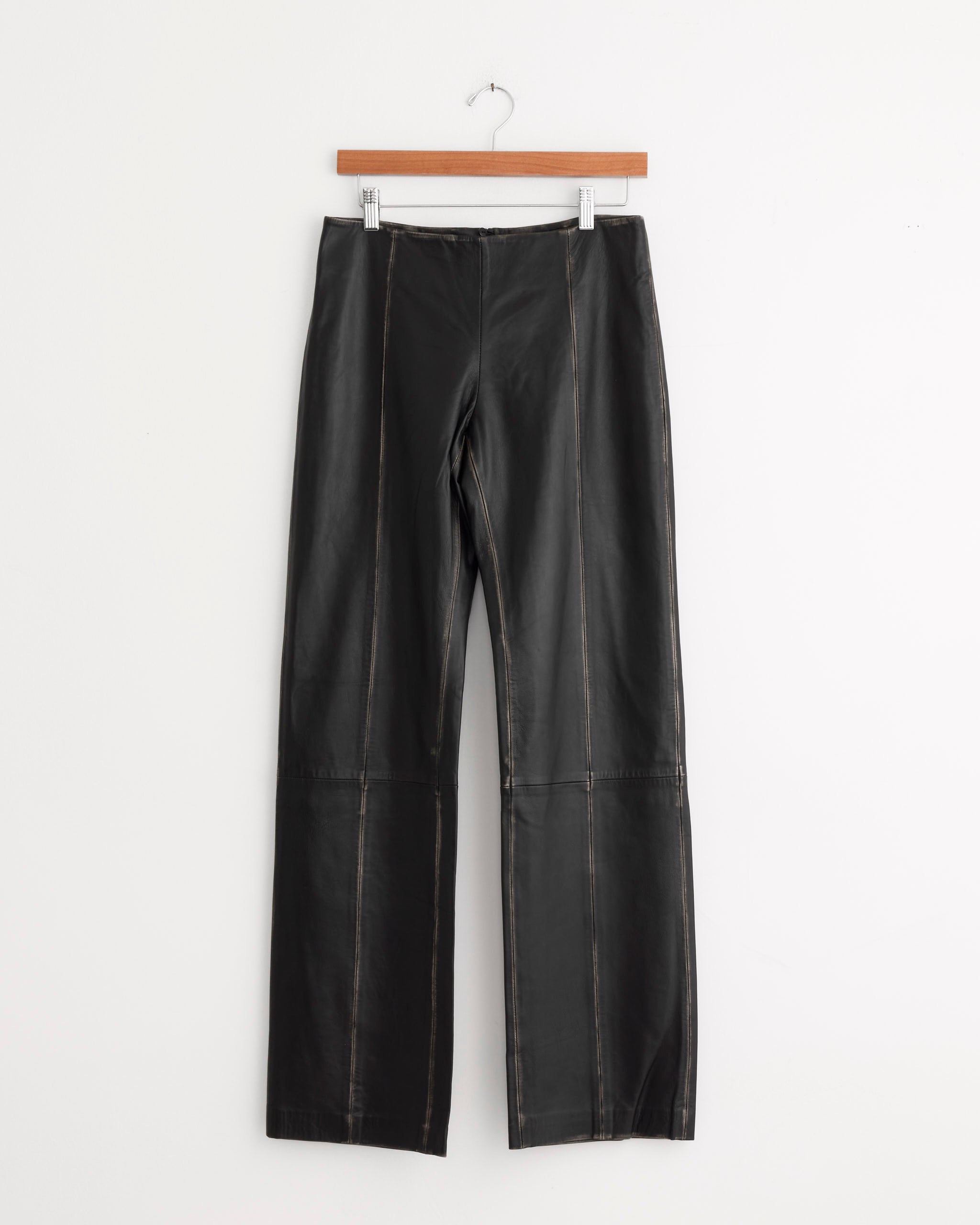 The Brushed Leather Pant in Black by ST. AGNI features visible seam detailing and a low-rise silhouette, crafted from black lambskin leather and displayed on a wooden hanger against a plain white background.