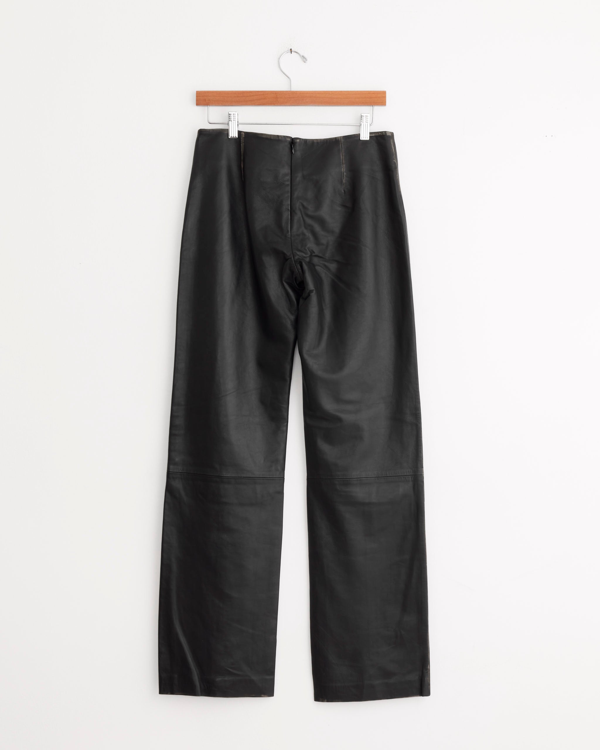 The ST. AGNI Brushed Leather Pant in Black, featuring a straight-leg cut and low-rise silhouette, hangs on a wooden hanger against a plain white background.
