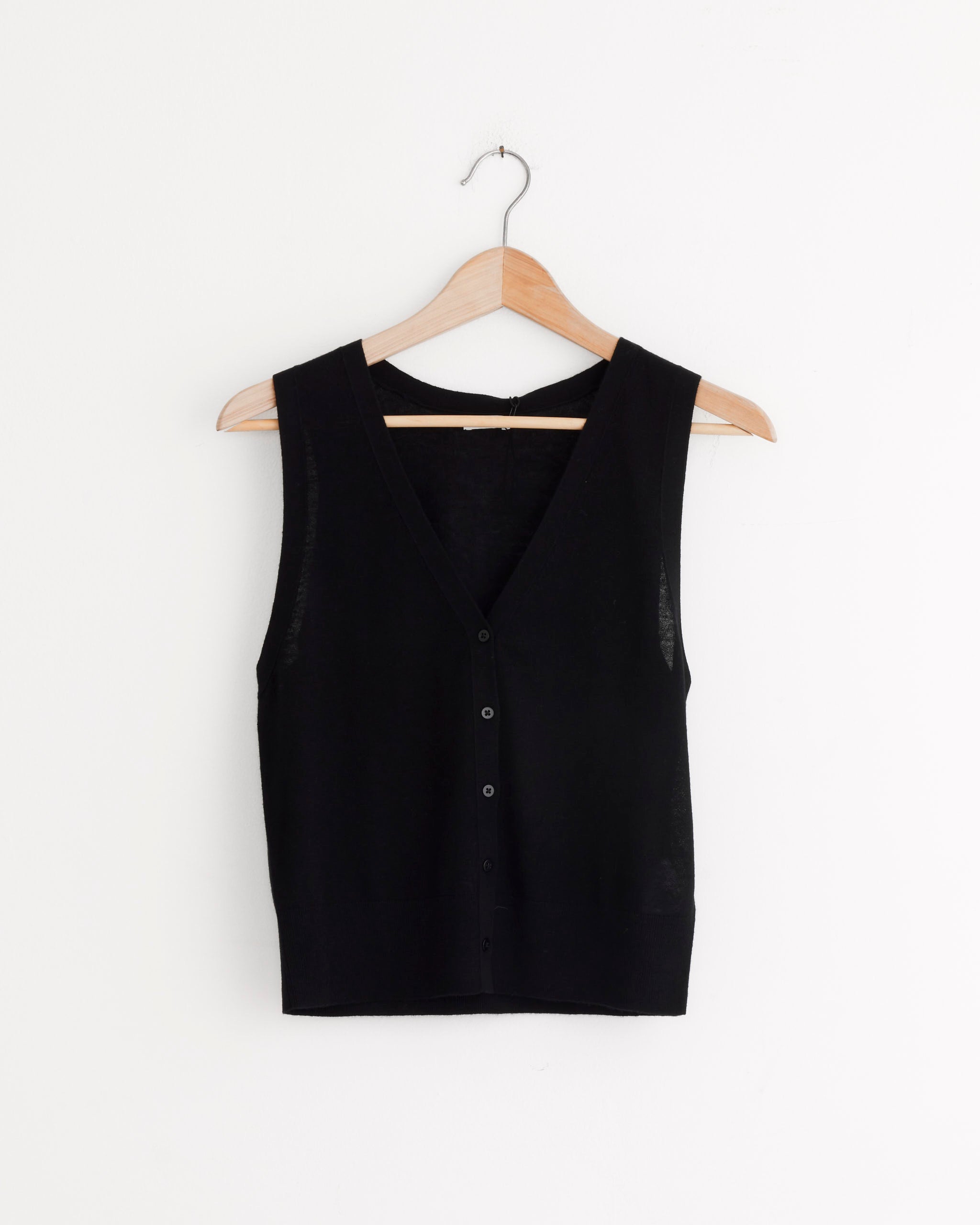 The Oupa Vest in Black by ST. AGNI is a sleeveless merino wool vest with a V-neckline and relaxed fit, shown on a wooden hanger against a plain white background.