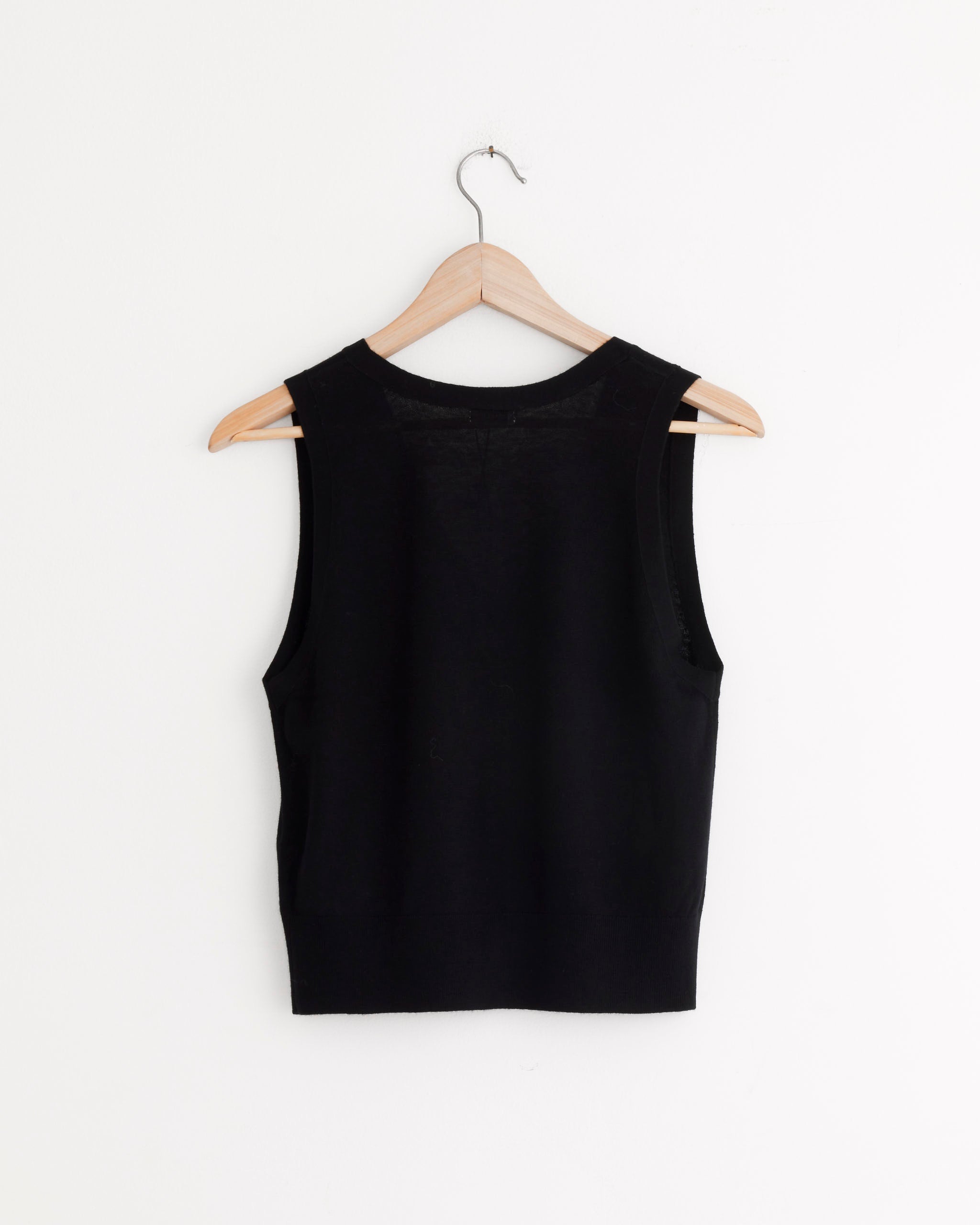 The Oupa Vest in Black by ST. AGNI, a sleeveless merino wool design with a relaxed fit, is displayed from the back on a wooden hanger against a plain white wall.