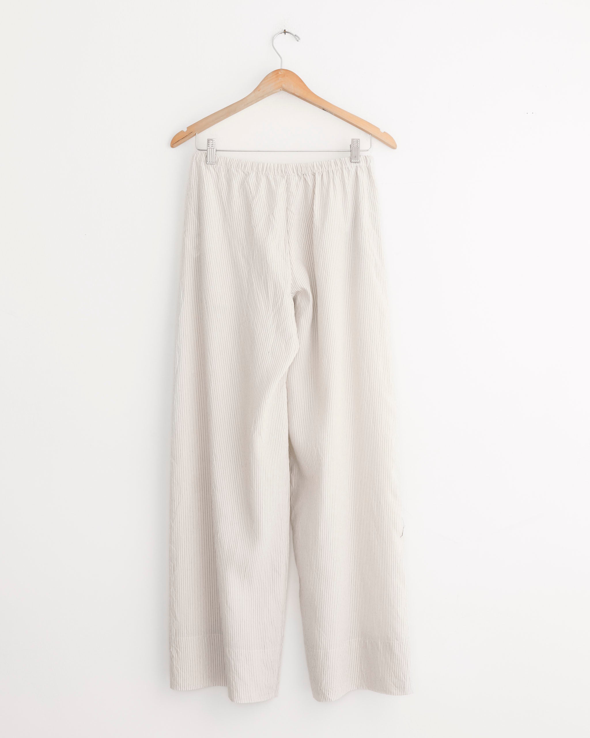 A pair of Relaxed Crushed Pant in Petite Pinstripe by ST. AGNI, featuring a mid-rise straight leg and elastic waistband, hangs on a wooden hanger against a plain white wall.