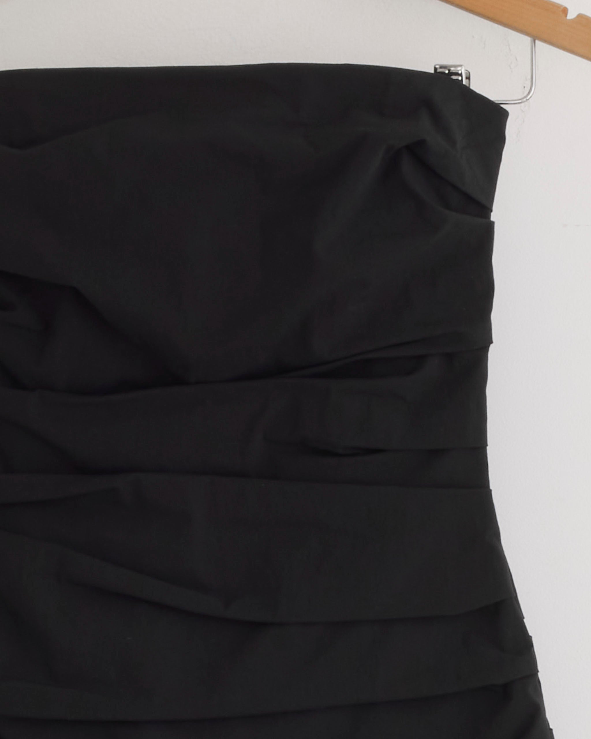 A close-up of the upper part of the Gathered Strapless Longline Dress in Black by ST. AGNI, featuring elegant ruching, hangs on a wooden hanger against a plain white wall.