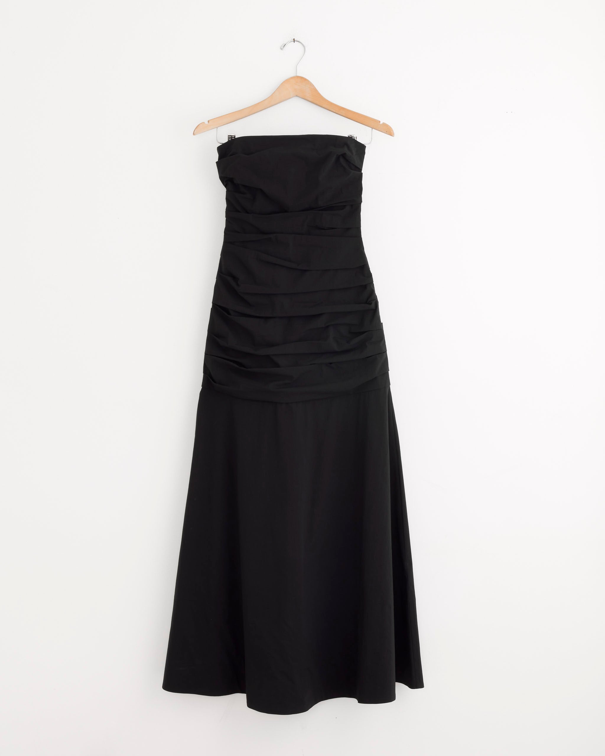 The ST. AGNI Gathered Strapless Longline Dress in Black, featuring a fitted ruched bodice and flared floor-length skirt, hangs elegantly on a wooden hanger against a plain white wall.