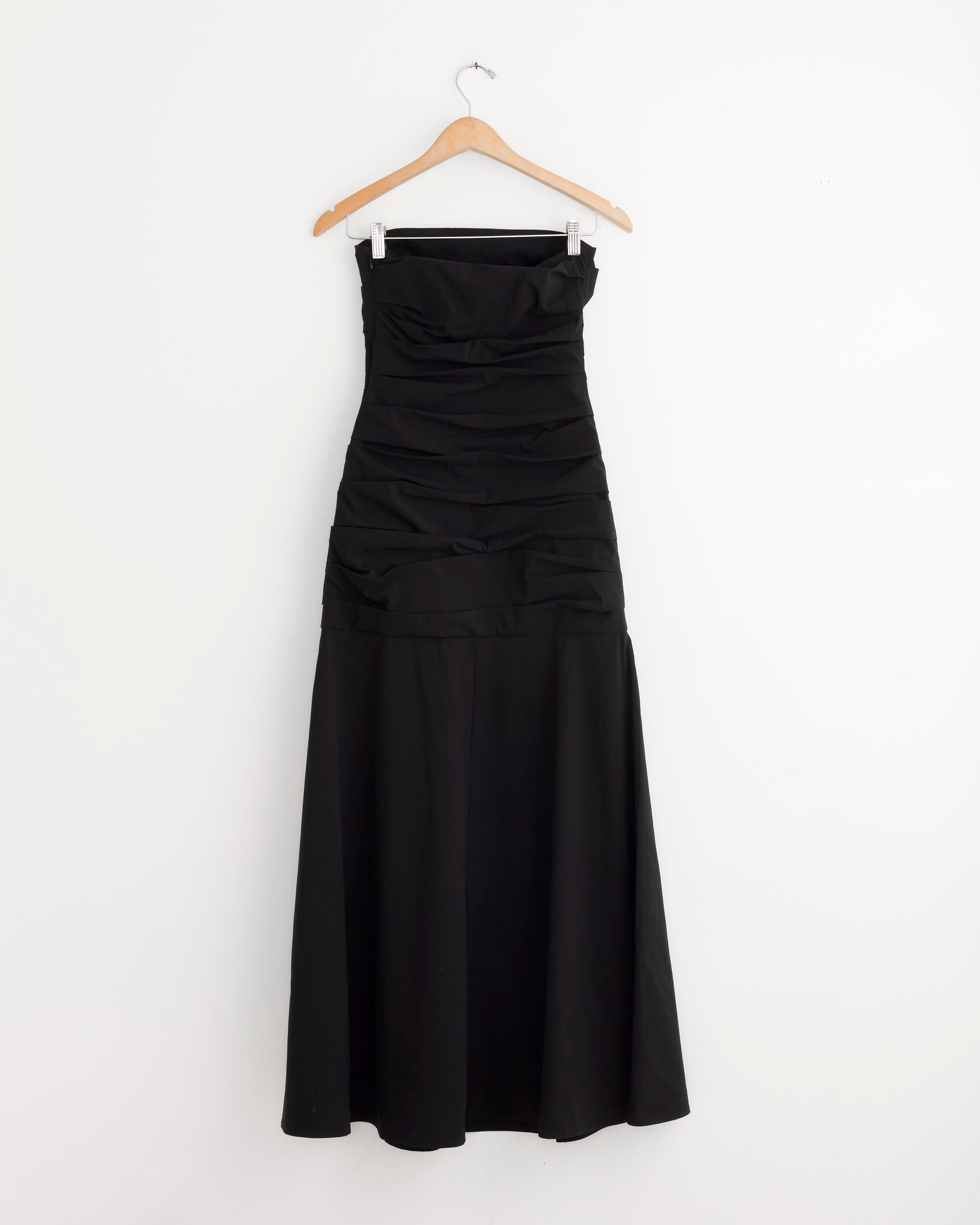 The ST. AGNI Gathered Strapless Longline Dress in Black features ruched bodice detailing and is displayed on a wooden hanger against a plain white wall.