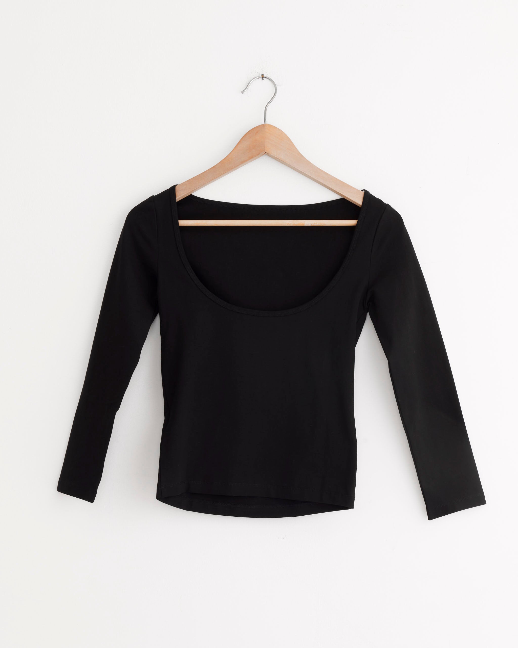 The ST. AGNI Ballerina Top in Jet Black, featuring long sleeves and a minimalist silhouette, hangs on a wooden hanger against a plain white wall.