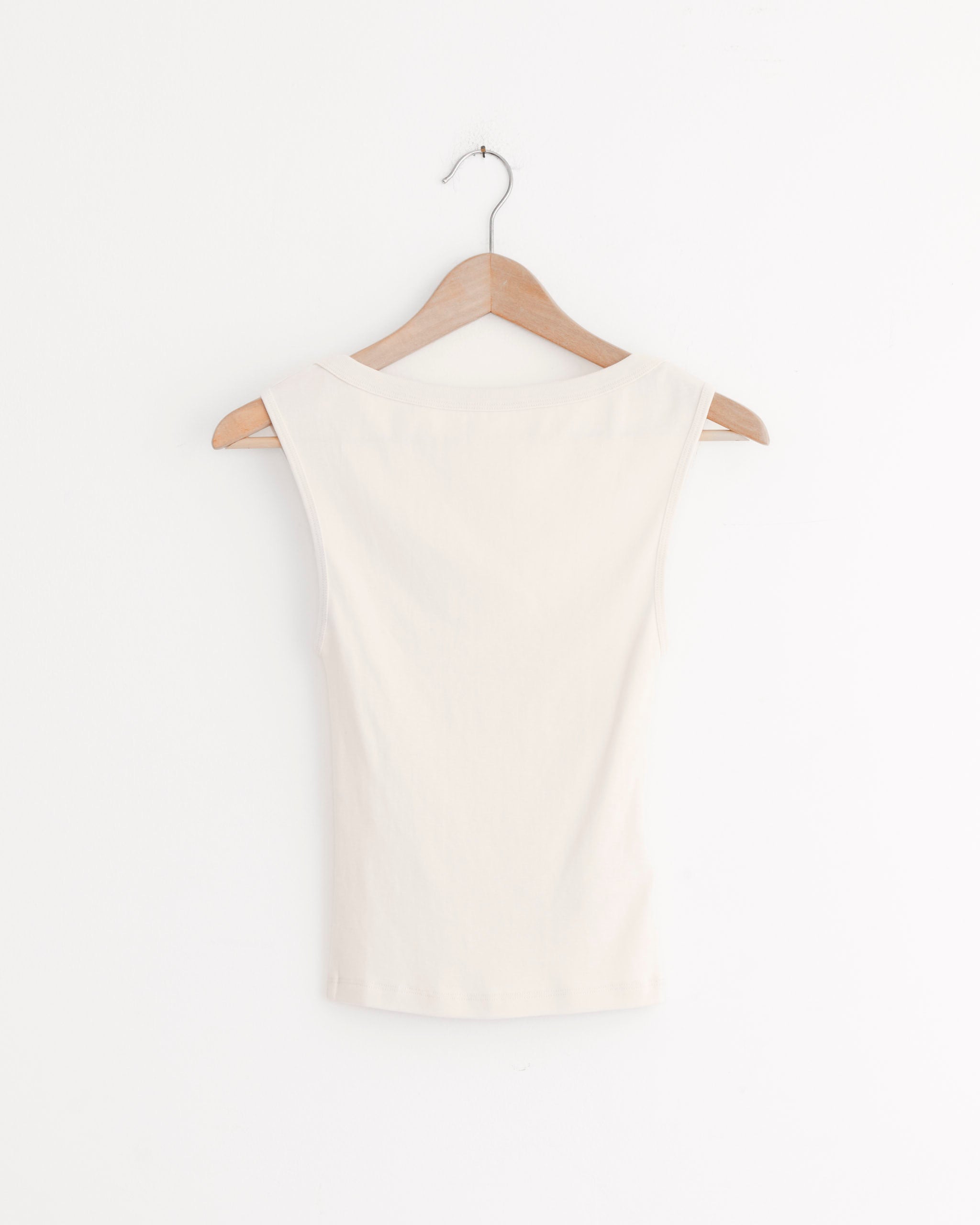 A sleeveless Ballerina Top in Shell Pink by ST. AGNI, crafted from organic cotton, is shown on a wooden hanger against a plain white wall with the back of the top facing forward.