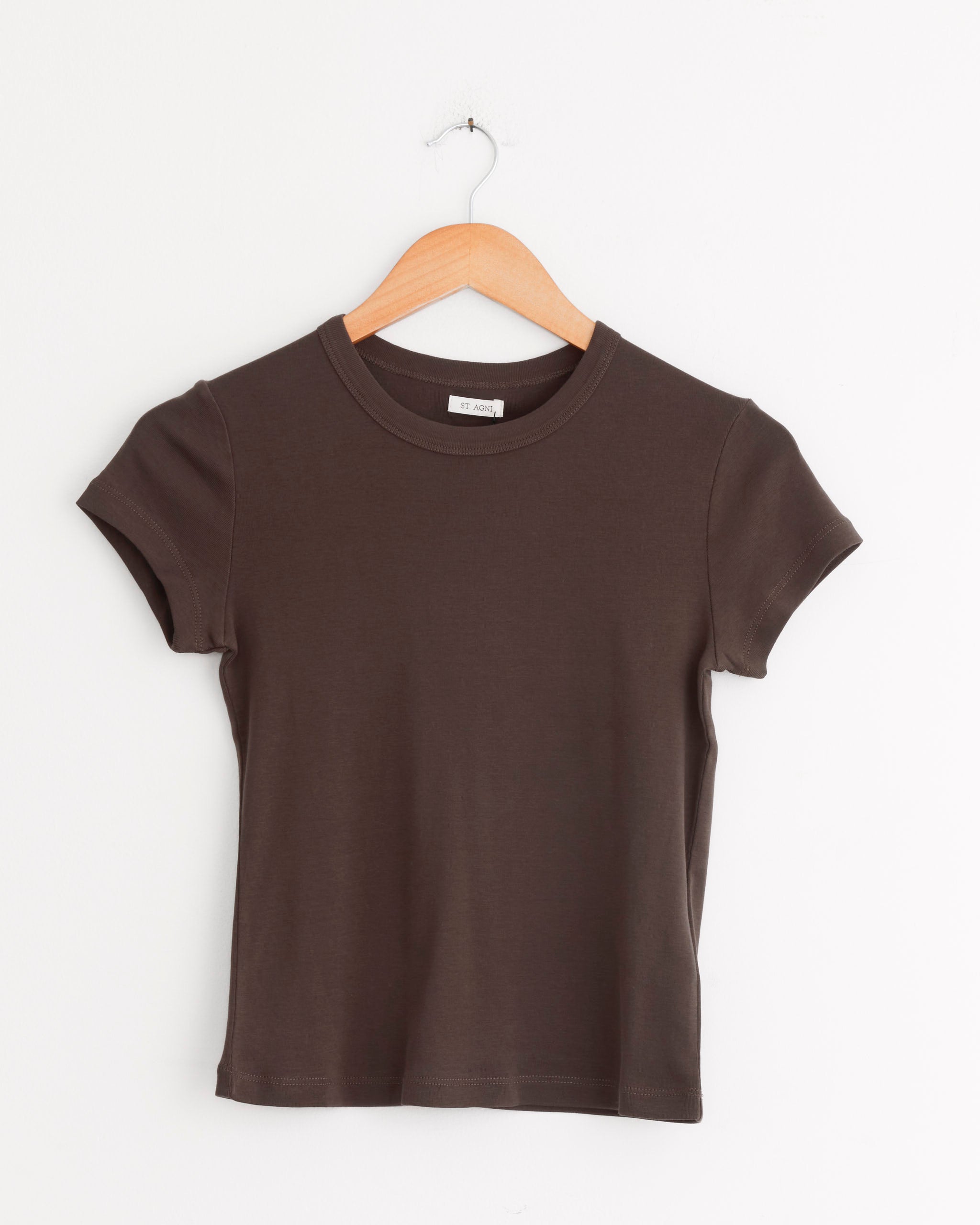 The ST. AGNI Baby Tee in Fudge, a dark brown short-sleeve top, hangs on a wooden hanger against a white wall.