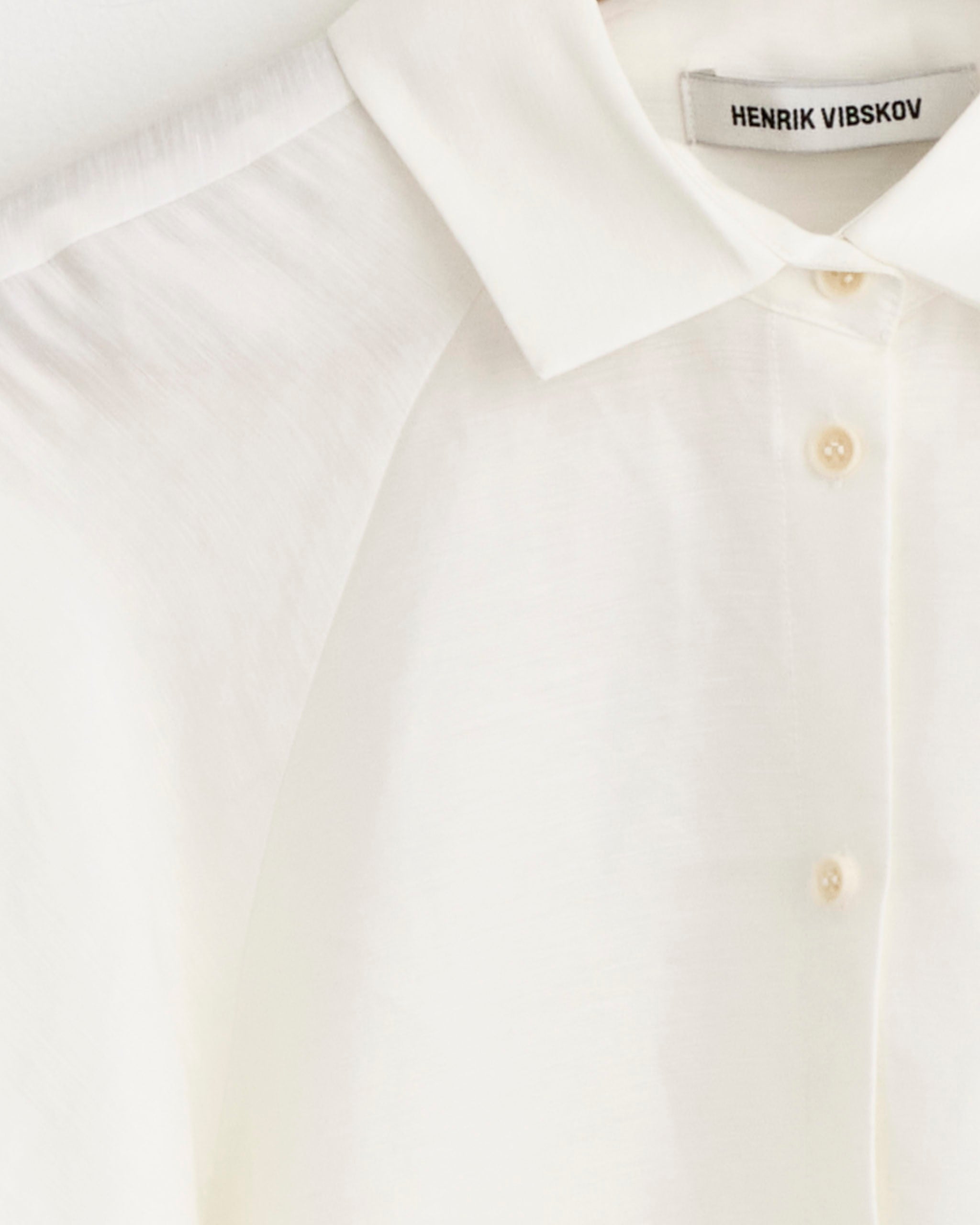 A close-up of the Case Shirt in White by Henrik Vibskov features cream buttons, the brand's label inside the neckline, and highlights the fit for accurate sizing.