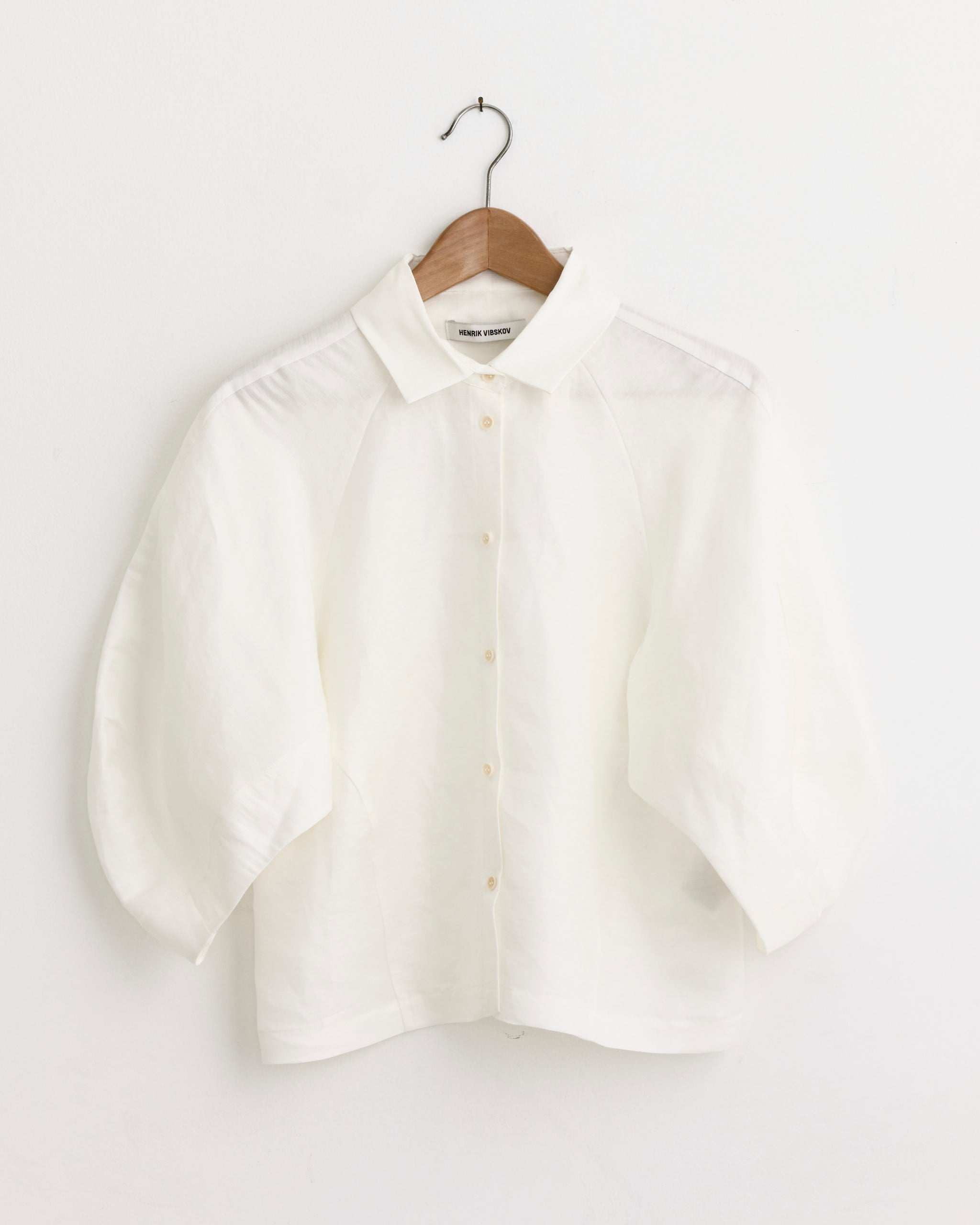 The Henrik Vibskov Case Shirt in White—a button-up blouse with a collar and wide three-quarter sleeves—hangs on a wooden hanger against a white wall. Chat online for fit and sizing questions.