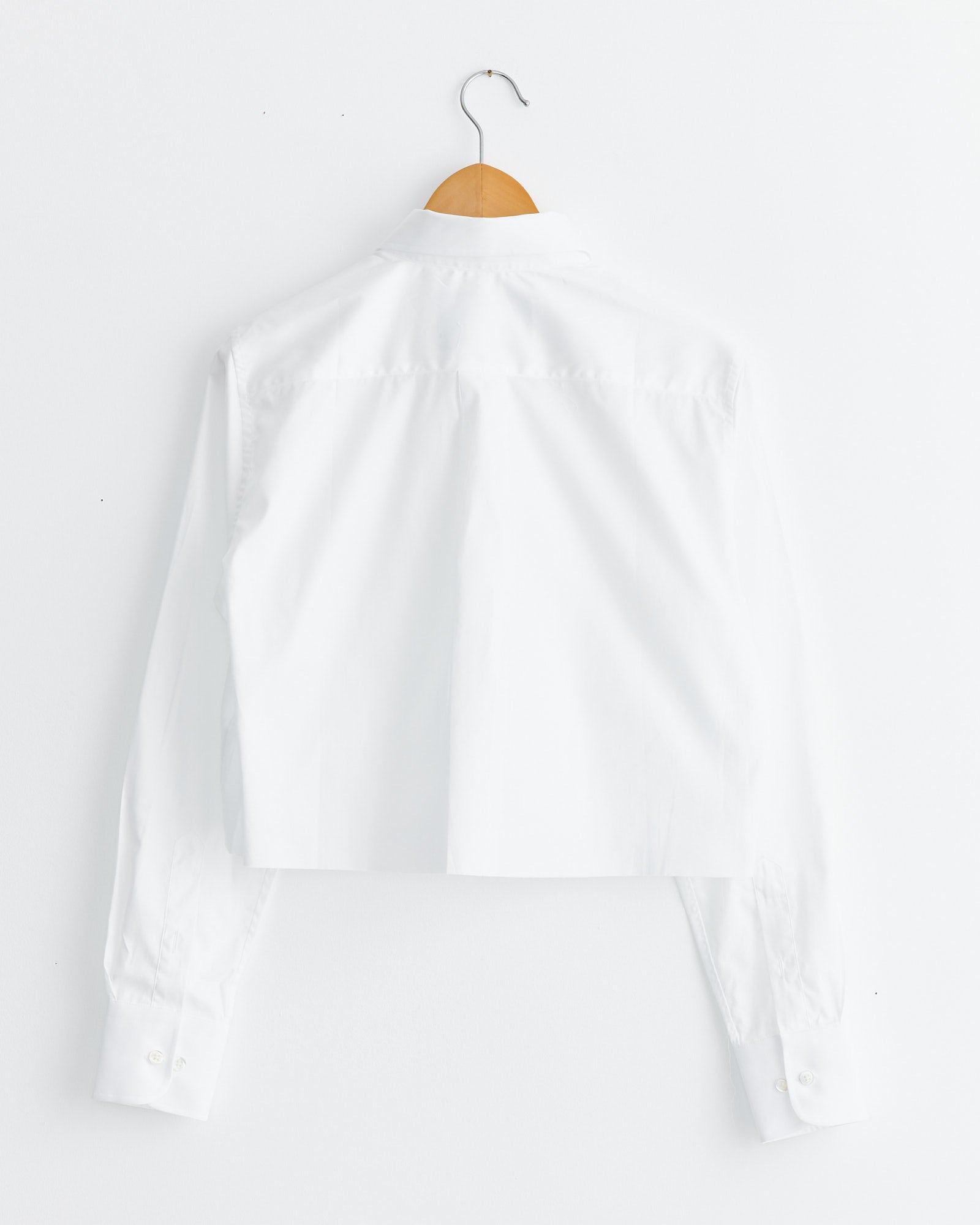 The Cropped Shirt in White by Hommegirls, shown from the back, hangs on a wooden hanger against a white wall. For fit or sizing questions, contact us via our online chat.