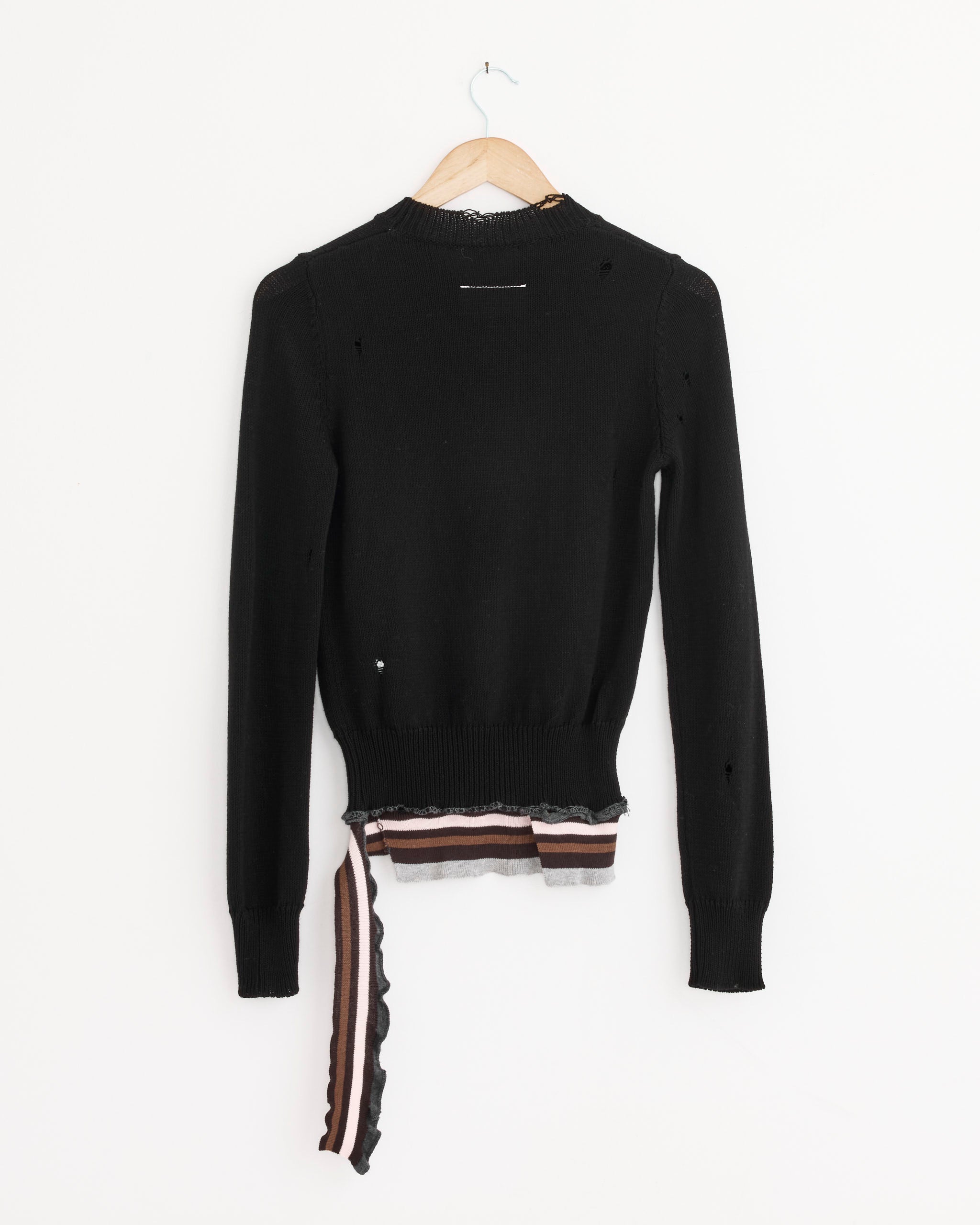 The MM6 Maison Margiela Crewneck in Black, a distressed wool sweater with ribbed silhouette, features small holes, signature stitch, and striped brown, black, and beige accents at the hem. Displayed on a wooden hanger against a white wall.