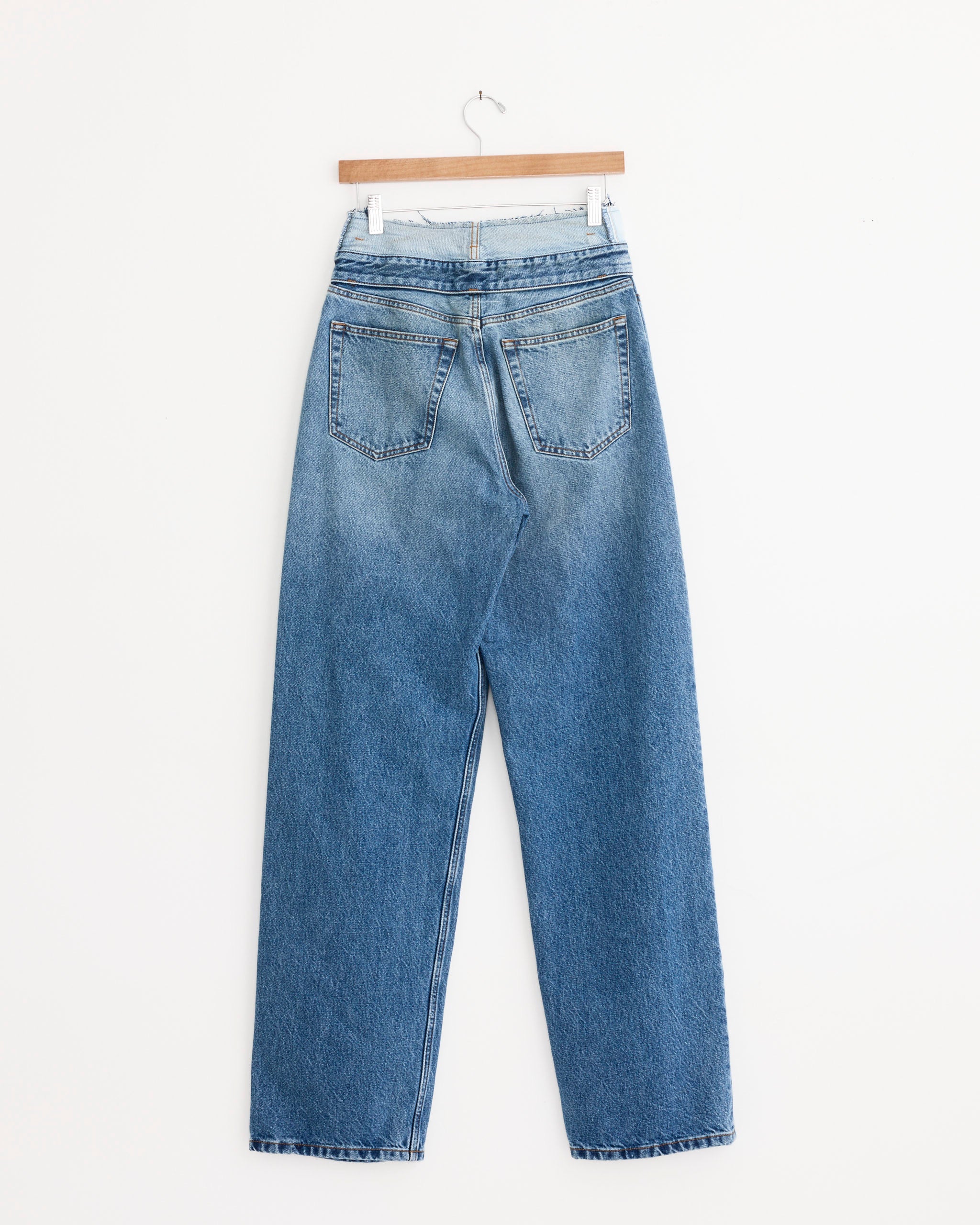 The Pockets Denim Jeans in Mid Blue by MM6 Maison Margiela, featuring a high-waisted straight-leg cut and back pockets, hang on a wooden hanger against a plain white wall.