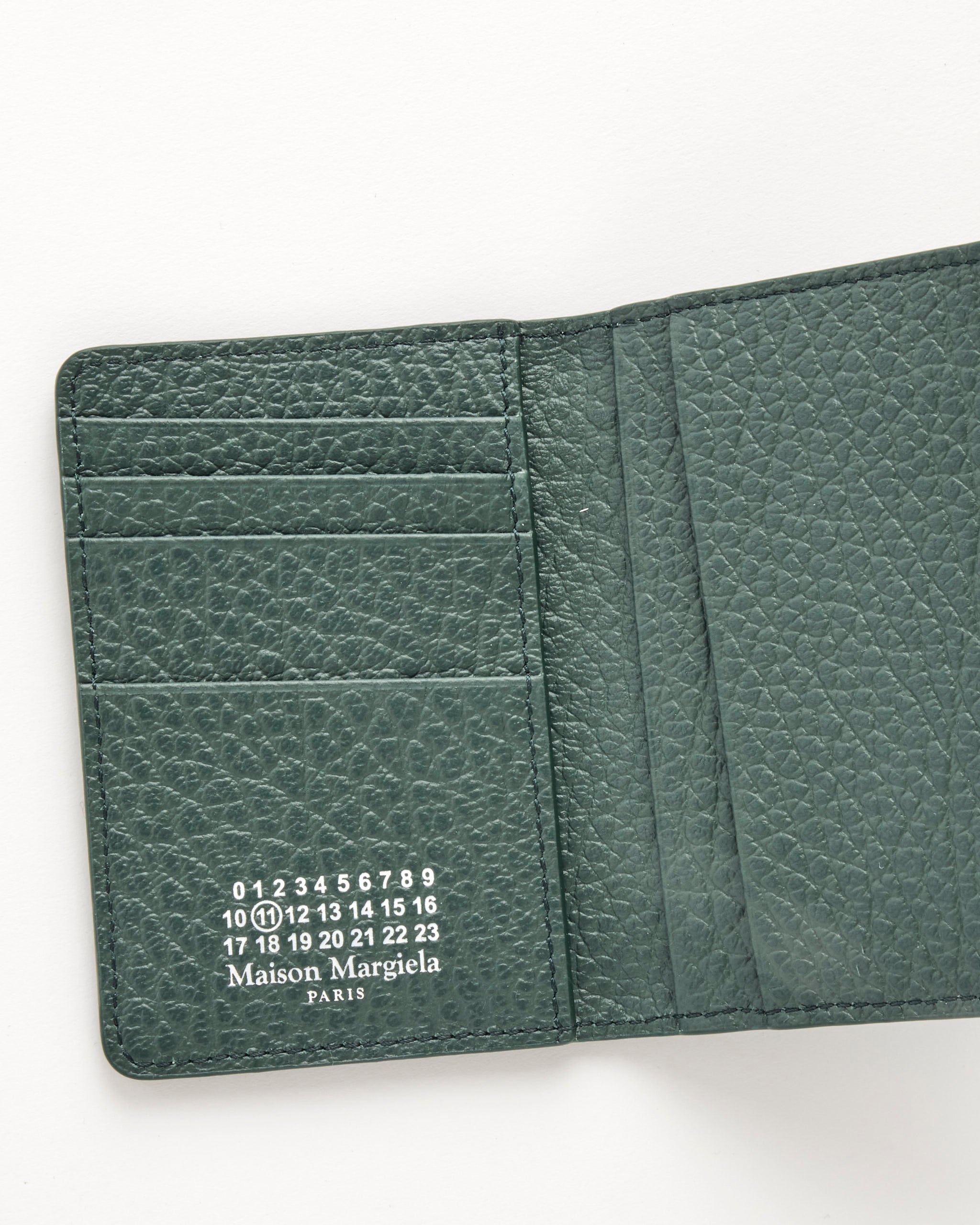 The Card Holder Slim 2 in Bottle Green by Maison Margiela is open on an off-white surface, revealing card slots and the brand's white-numbered logo printed inside on textured, embossed green leather.