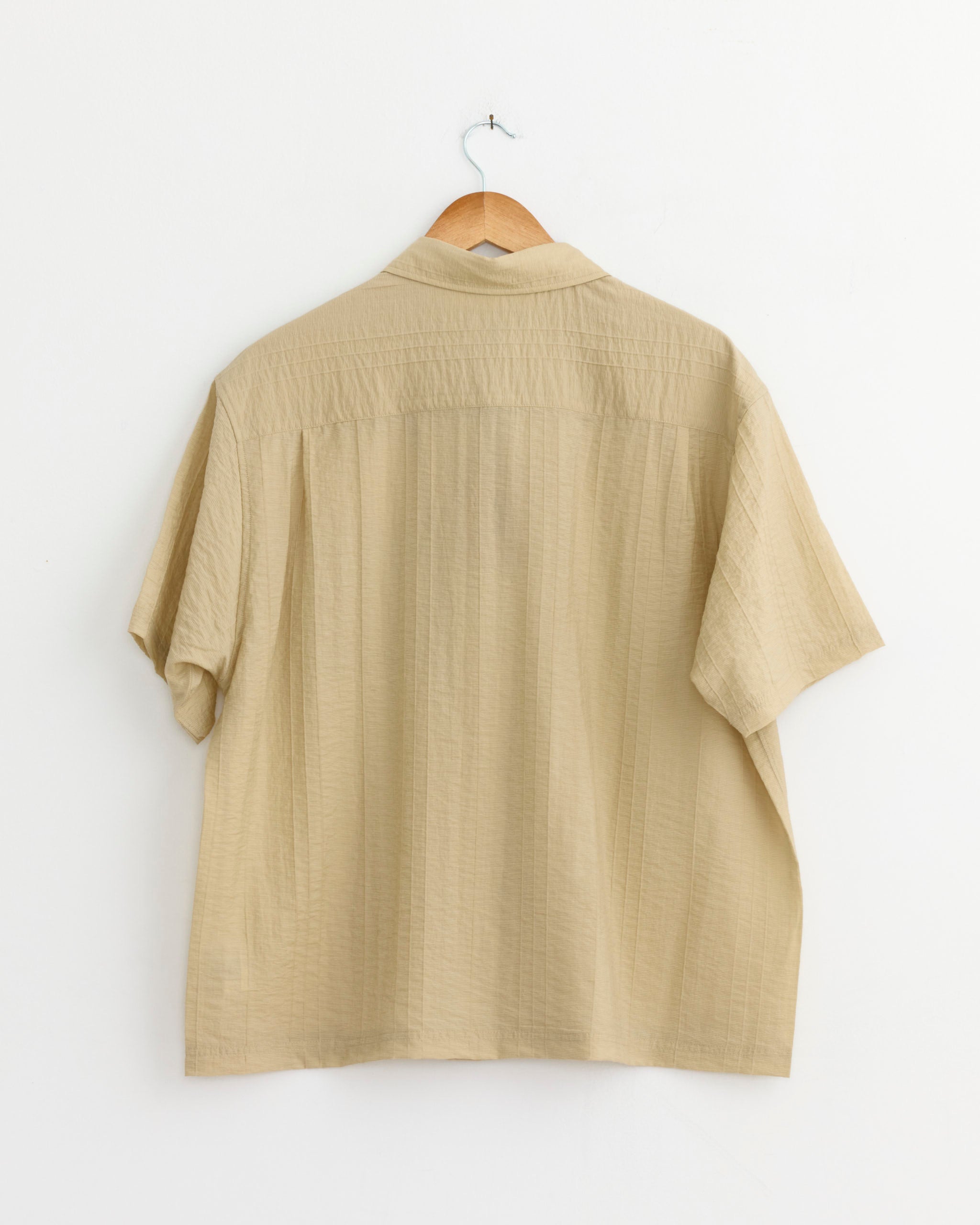 The Stussy Pleated Textured Shirt in Tan hangs on a wooden hanger against a white wall, shown from the back. This unisex shirt features short sleeves, a loose fit, and subtle textured detailing.