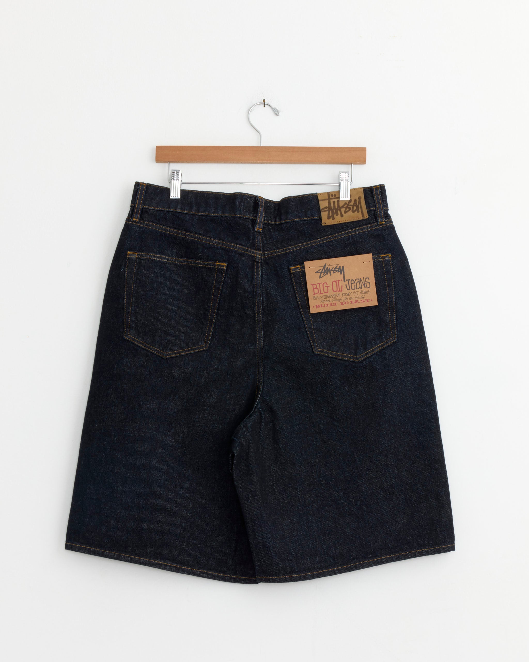 Big Ol Denim Short in Rinsed Indigo by Stussy, shown from the back on a wooden hanger. These heavyweight cotton shorts feature two back pockets: one with a tan Stussy label, the other with a small brown waistband label.