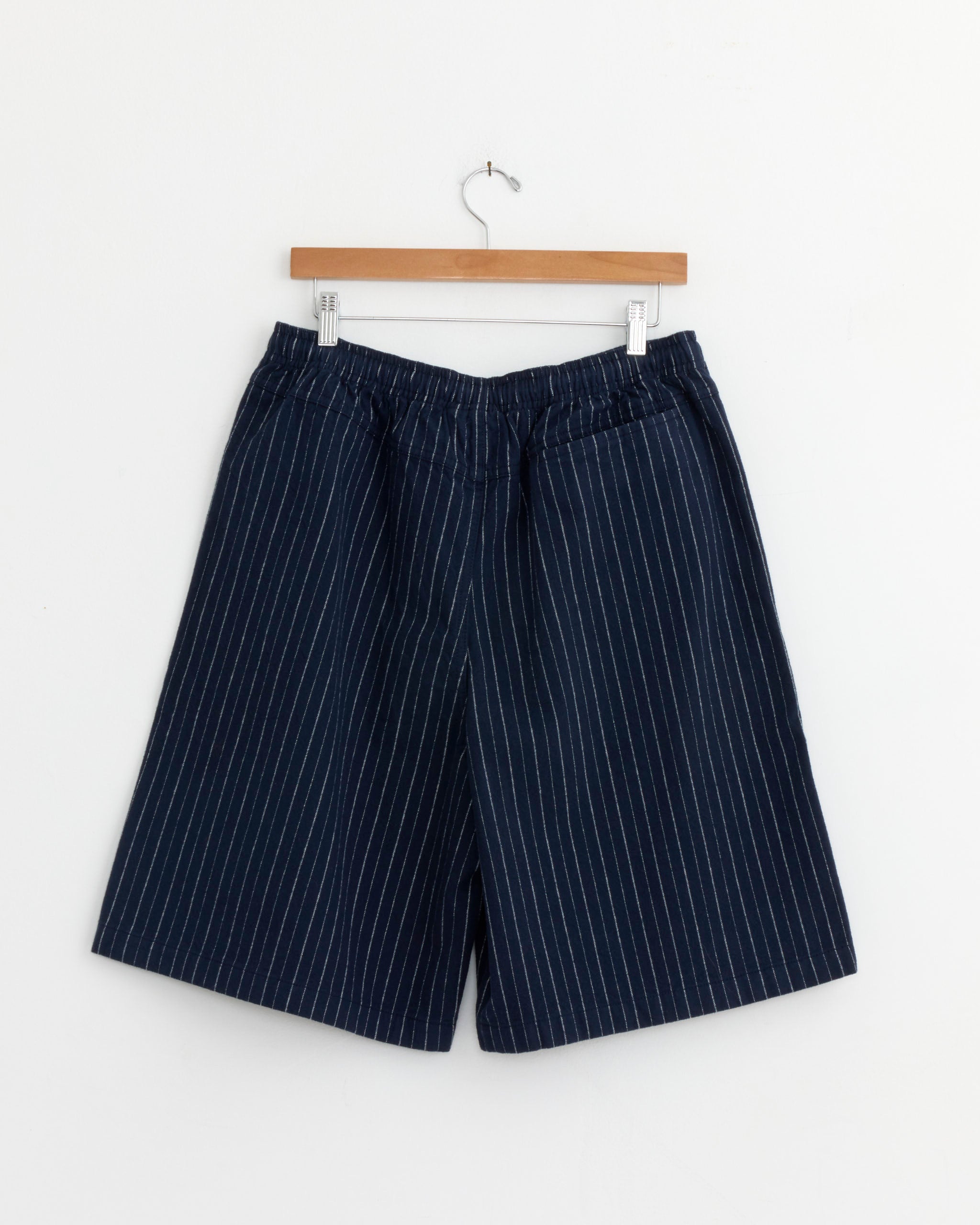Stussy Baggy Beach Short in Navy with white vertical pinstripes and an elastic waistband, displayed on a wooden hanger against a white background.