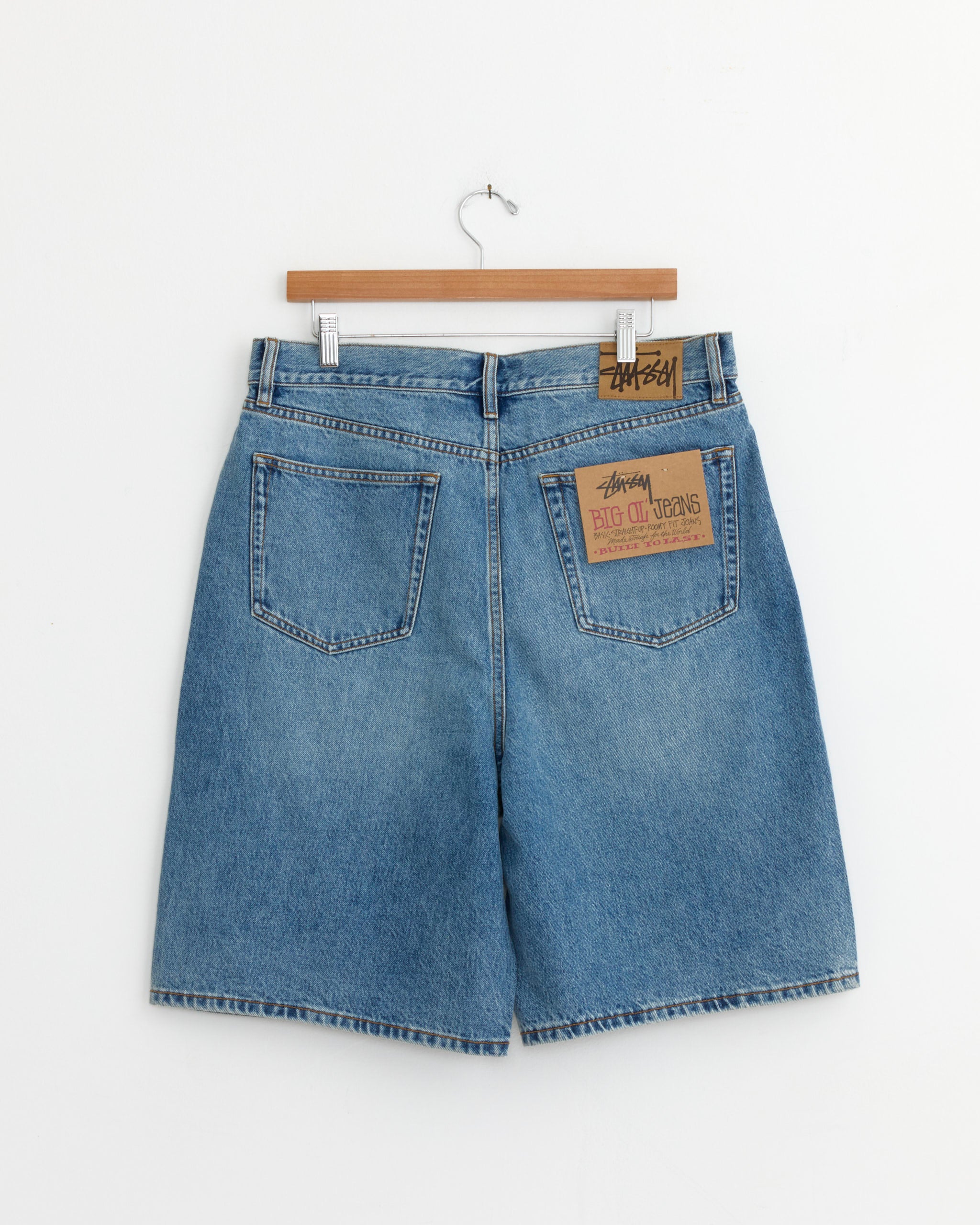 The Stussy Big Ol Denim Short in Indigo Stone Wash is shown from the back on a wooden hanger. It has two back pockets, a brown leather "Stussy Big Ol Jeans" patch above the right pocket, and a relaxed, baggy fit.