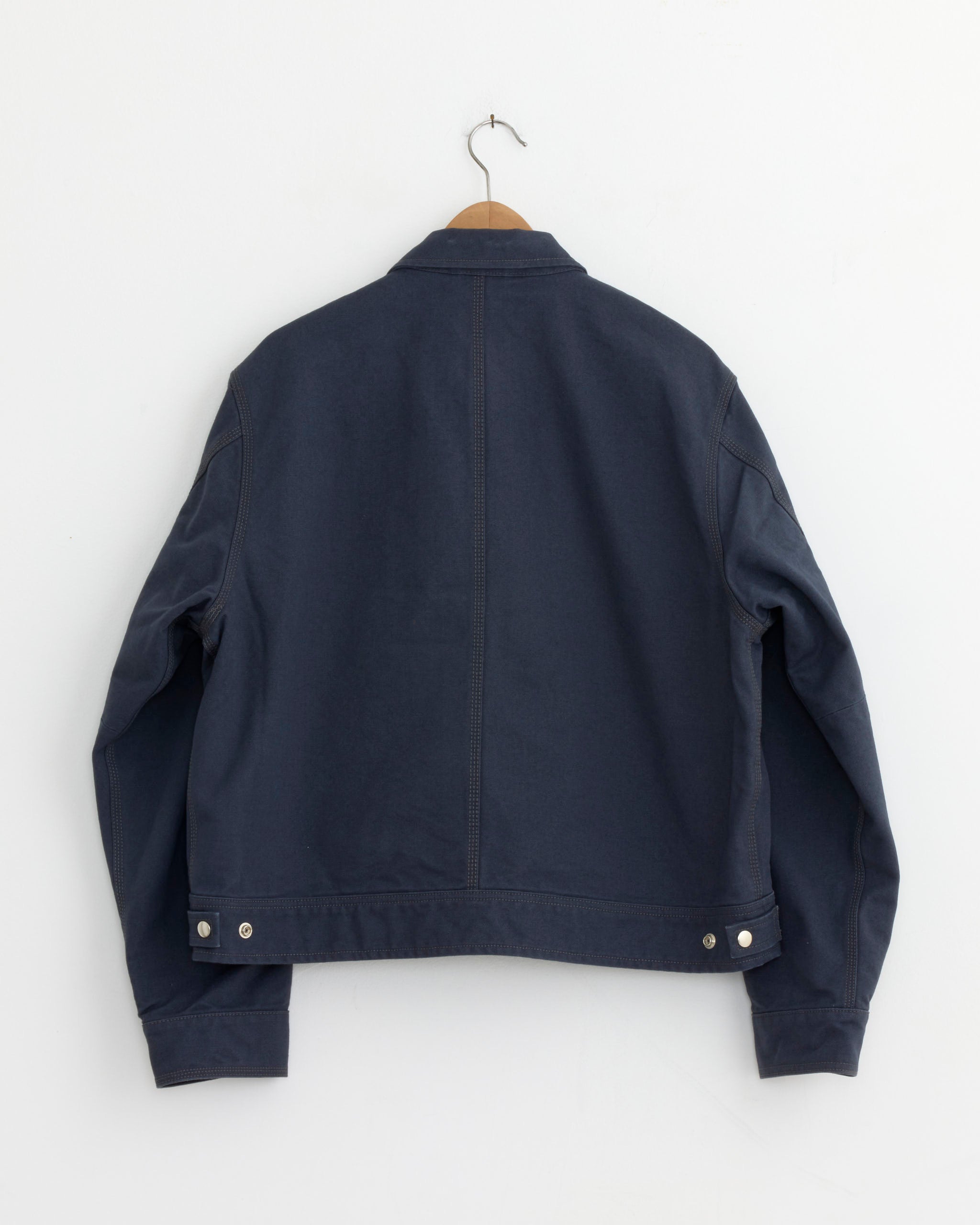 The Work Collared Jacket in Blue by Stussy, made from cotton canvas, hangs on a wooden hanger against a white wall. Displayed from the back, it has long sleeves and silver buttons at the waist and cuffs.