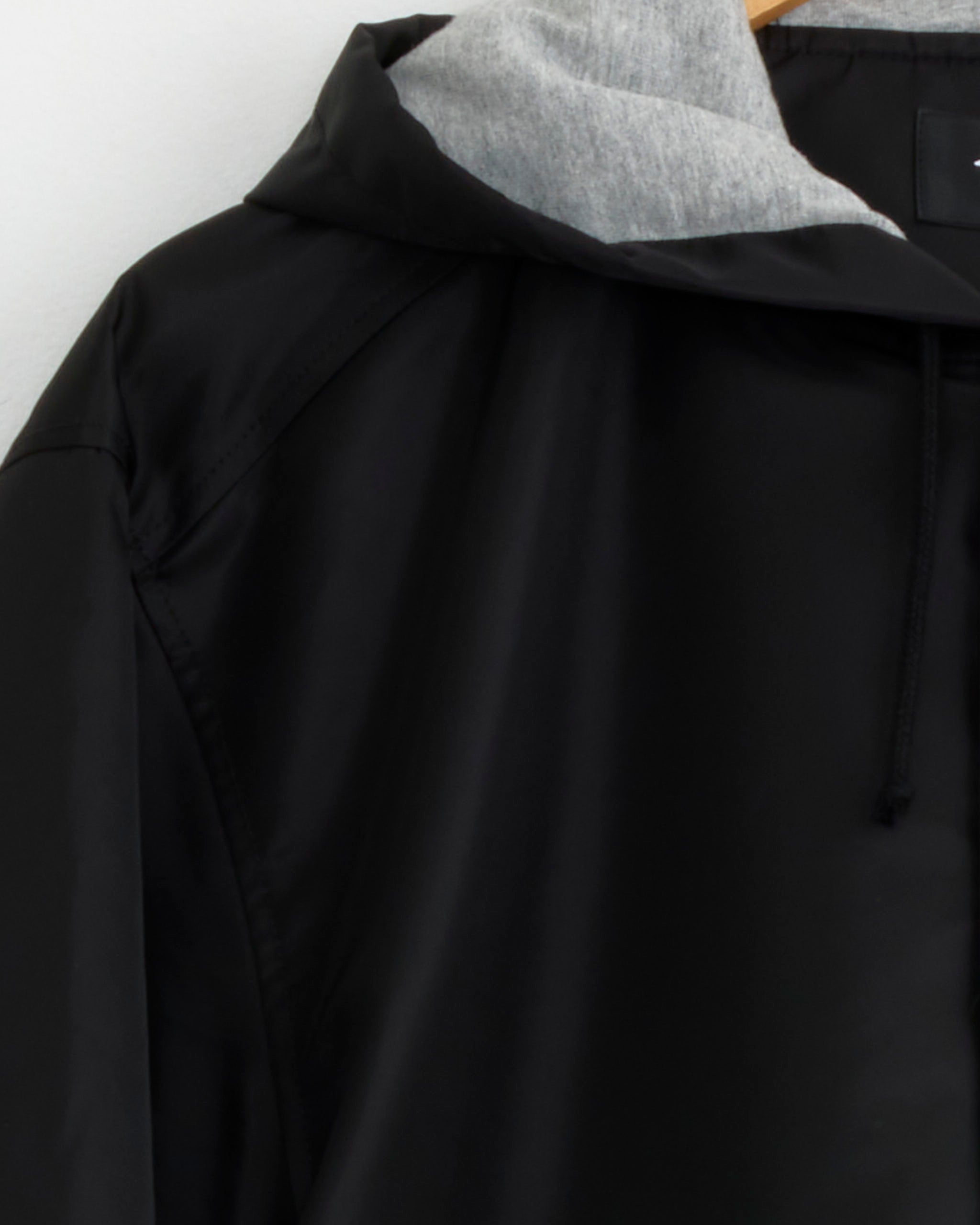 A close-up of the Stussy Hooded Coaches Jacket in Black, featuring a gray hood, displayed on a wooden hanger against a white wall.