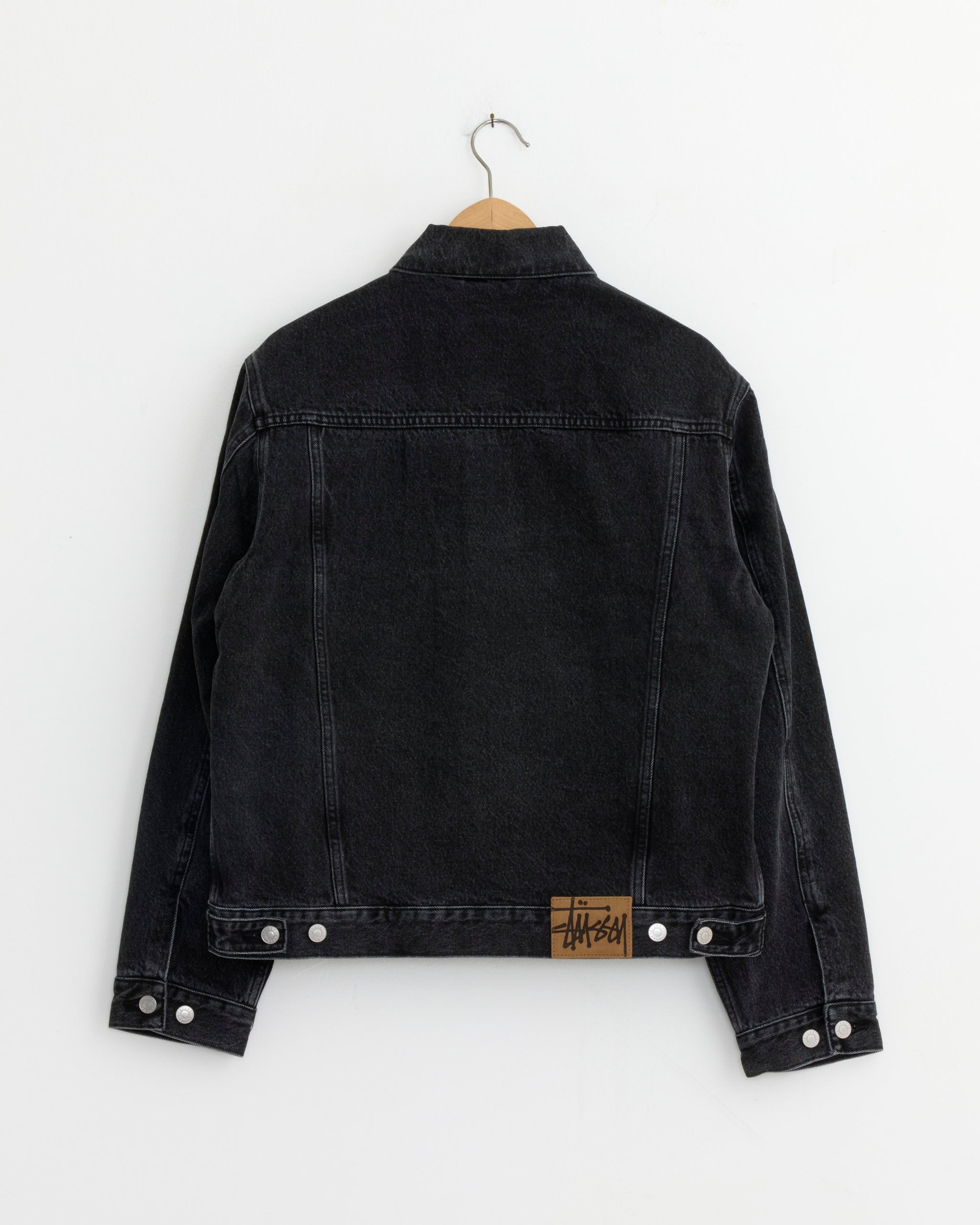 The Stussy Lined Denim Trucker Jacket in Vintage Black hangs on a wooden hanger against a white wall, shown from the back. A tan leather patch with black text is sewn near the right bottom hem.