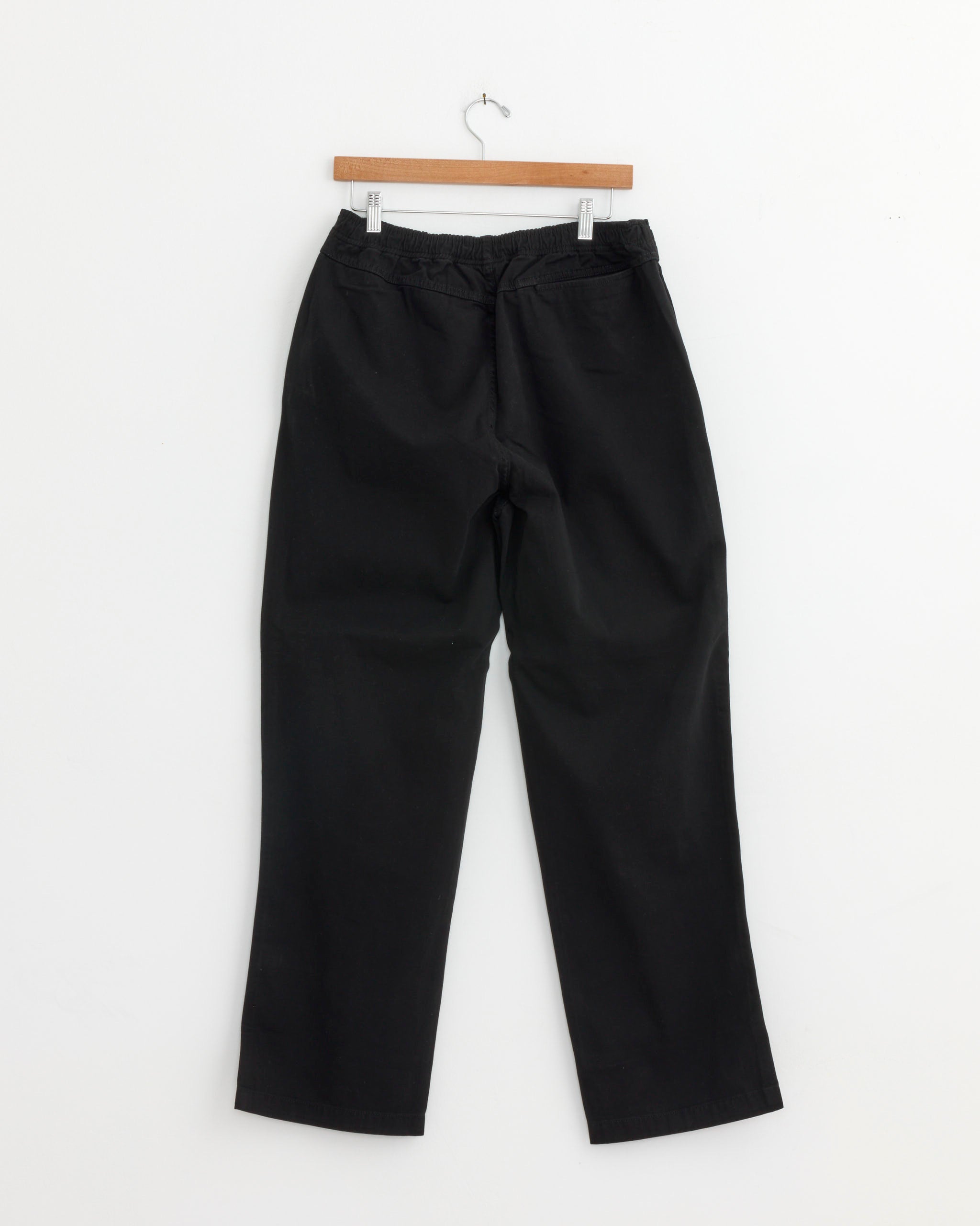 The Stussy Beach Pant in Black, featuring a unisex relaxed fit and cotton twill fabric, hangs on a wooden hanger against a plain white wall, displayed from the back.