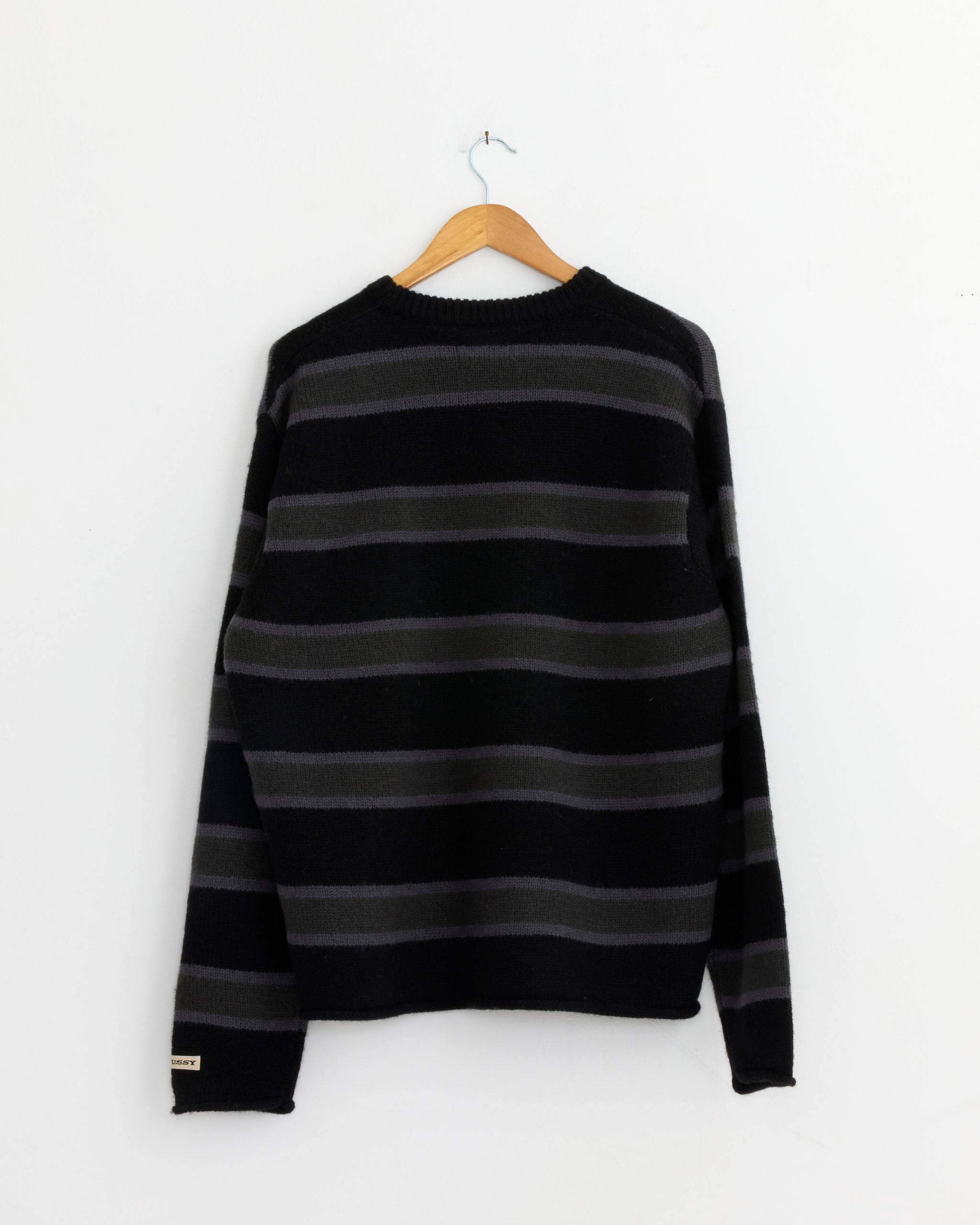 The Stussy Striped Wool Sweater in Black hangs on a wooden hanger against a white wall.