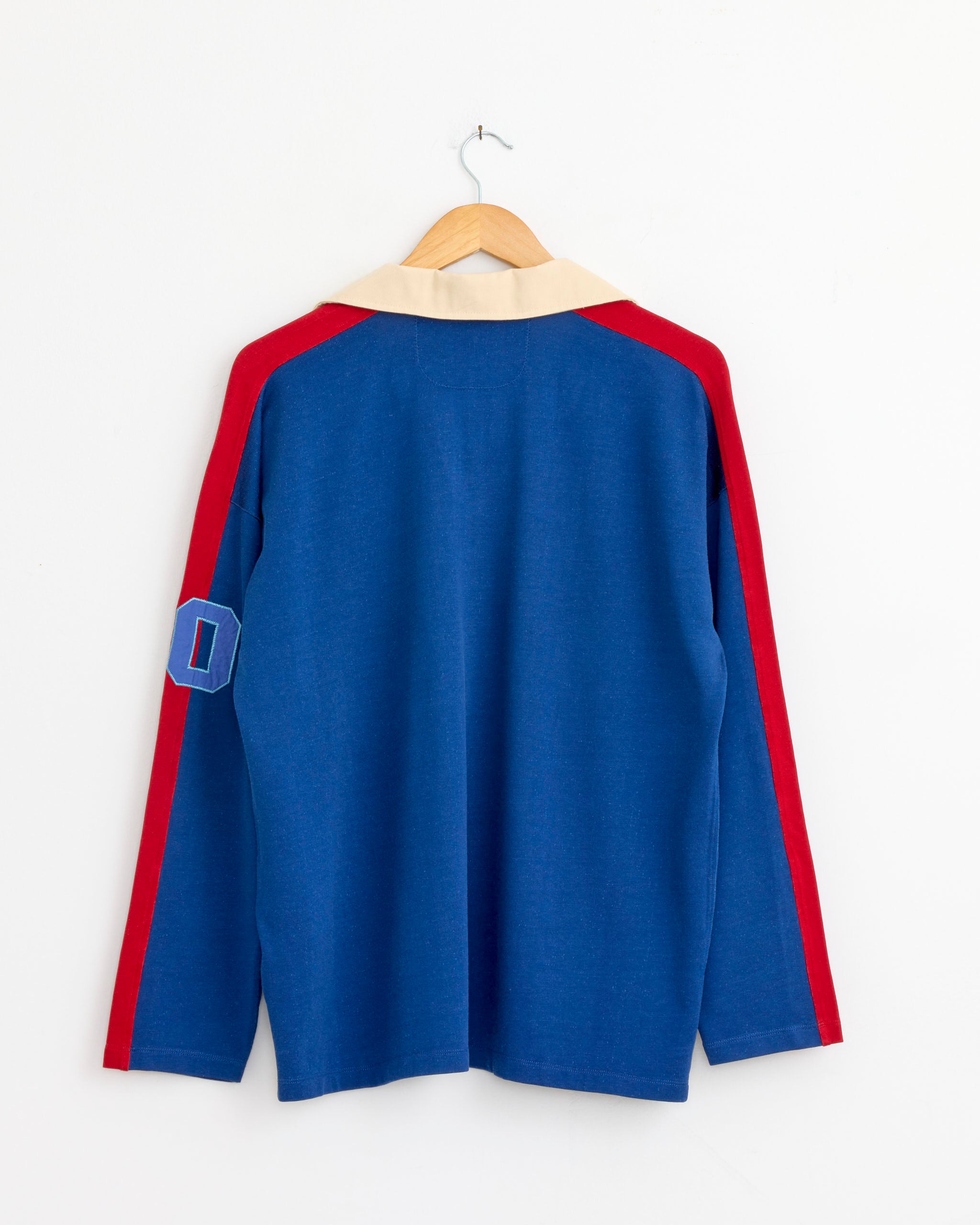 The Bode Lakewood Polo in Blue features long sleeves with red stripes, a beige collar, and a blue patch with a red border and white "D" on the left arm, made from Portuguese fabric. It’s shown on a wooden hanger against a white wall.