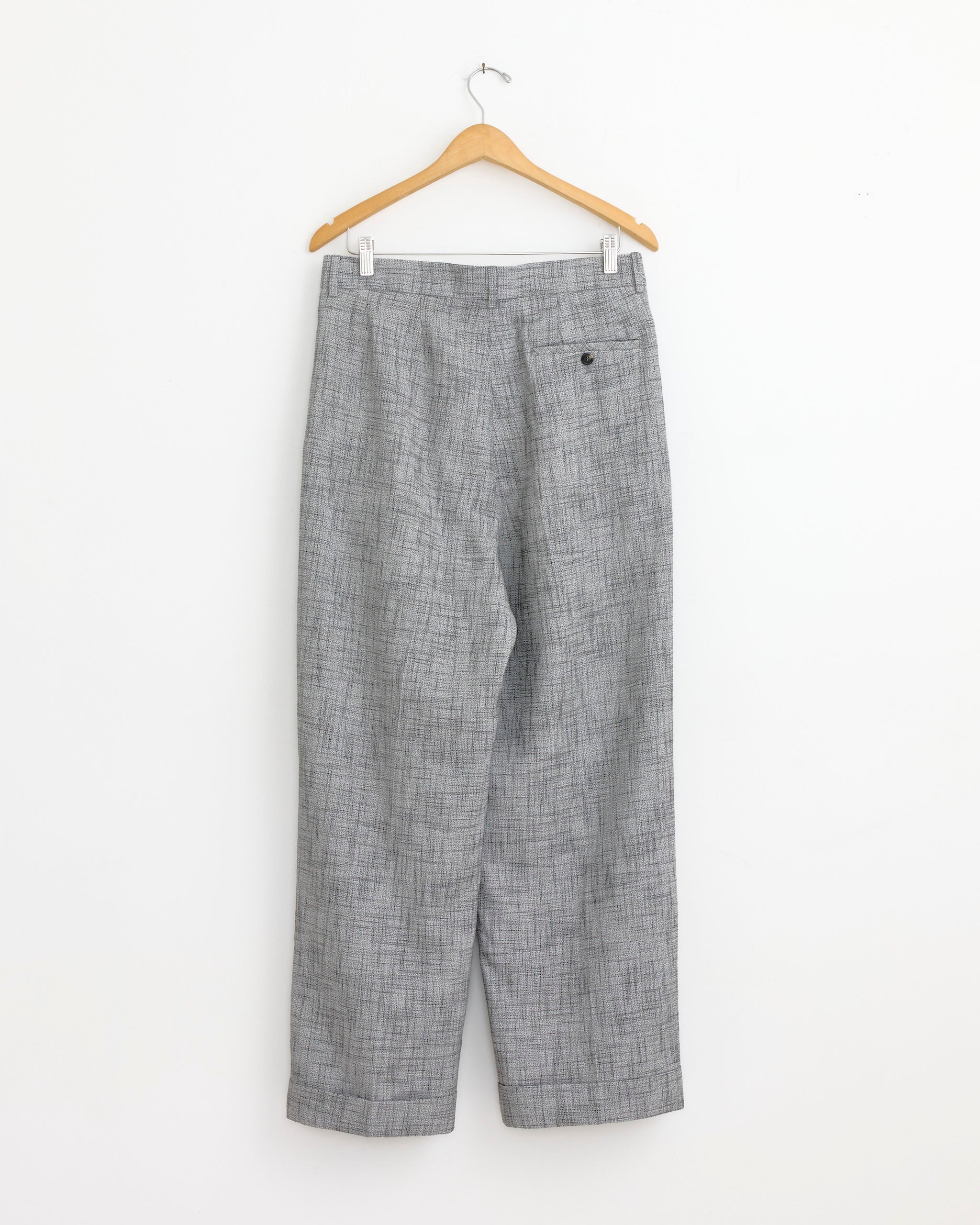 The Bode Static Weave McNab Trouser in Grey, featuring a subtle plaid pattern and wide-leg design, hangs on a wooden hanger against a plain white wall.