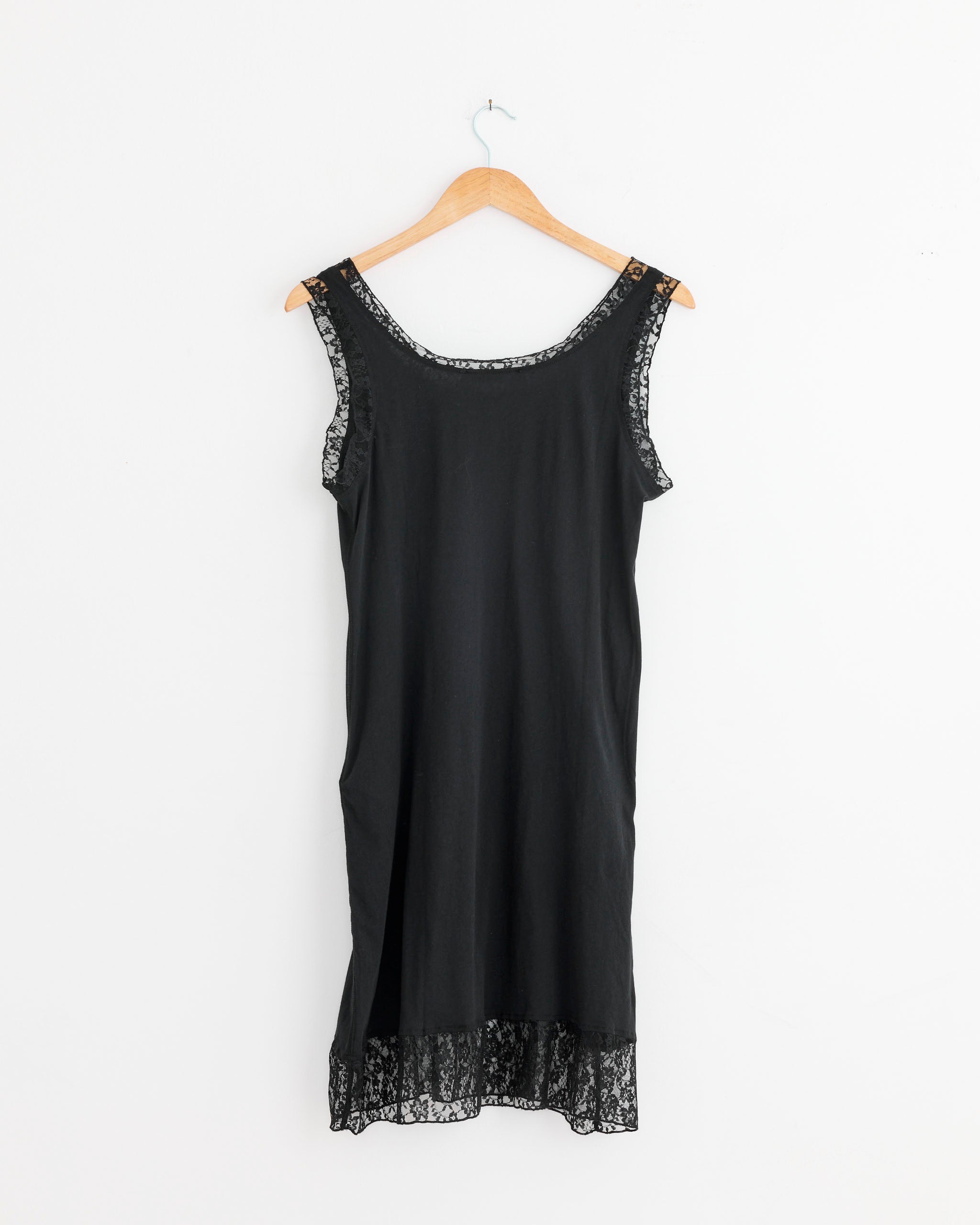 The Our Legacy Dawn Dress in Raven Mist, a sleeveless black dress made from Airveil Cotton Portuguese jersey with lace trim at the neckline, shoulders, and hem, is displayed on a wooden hanger against a plain white wall.