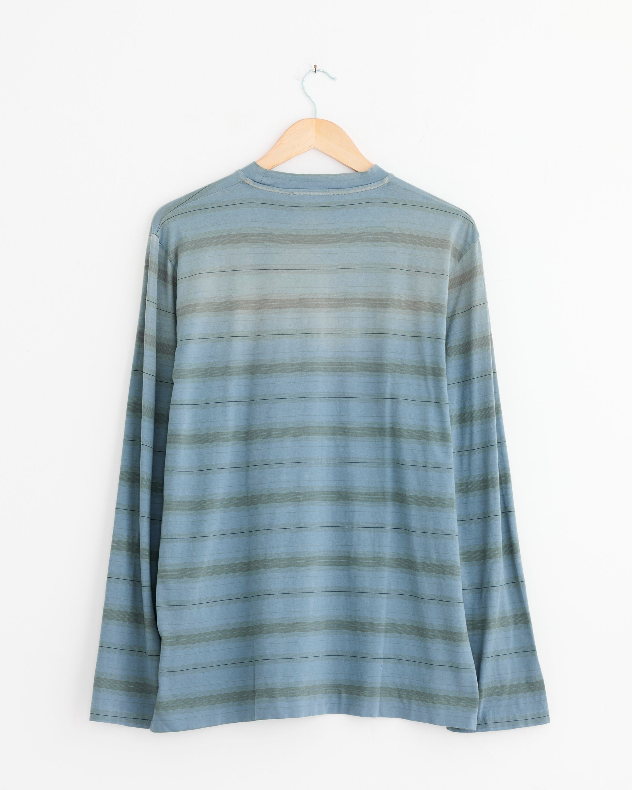 The Box T-Shirt in Spray Fade Ghost Stripe by Our Legacy, featuring long sleeves and blue-gray stripes, hangs on a wooden hanger against a white wall—ideal for classic style lovers. For fit or sizing help, use our online chat.