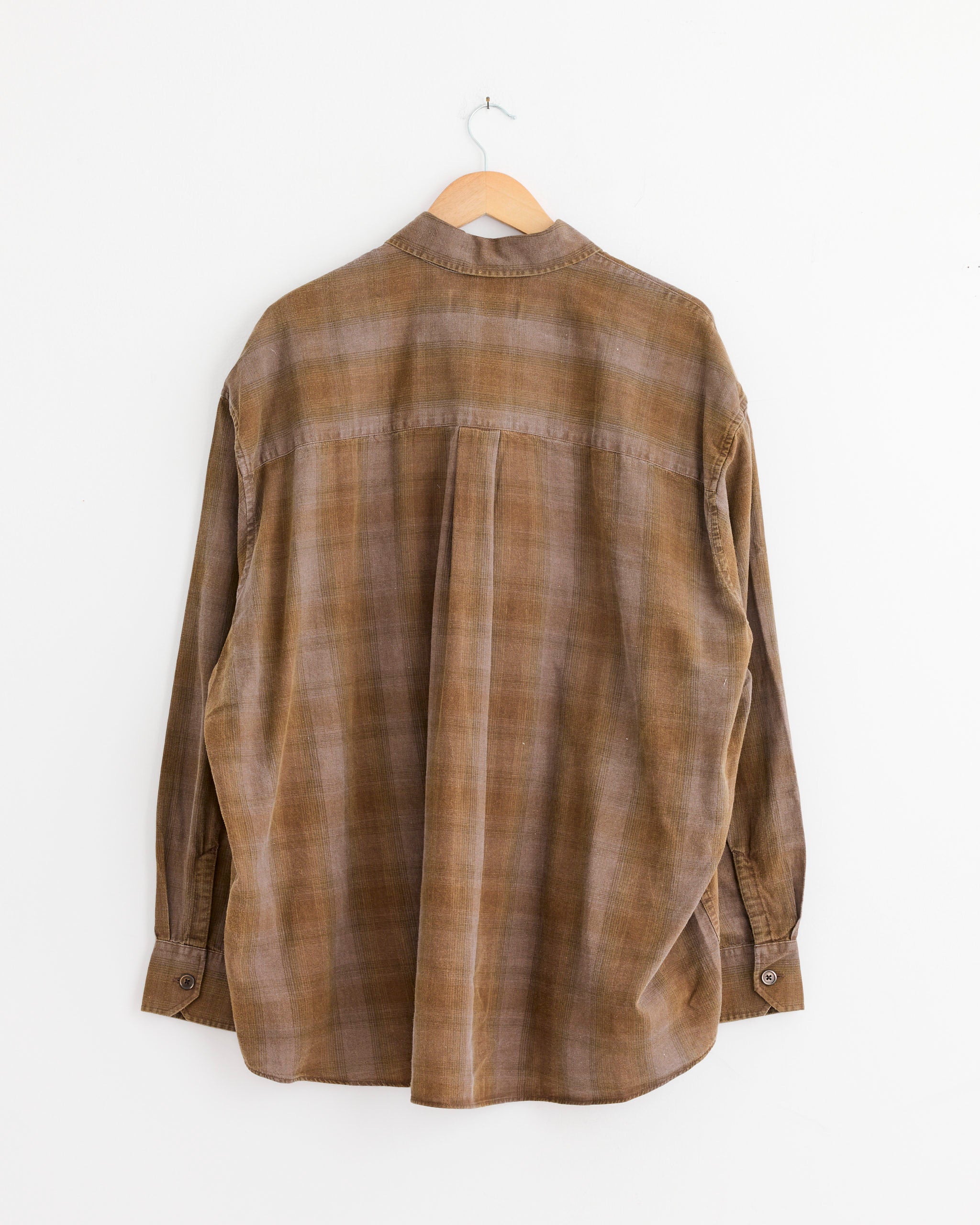 The Borrowed Shirt in Dakota Flannel Check by Our Legacy, crafted from Portuguese cotton flannel, is displayed hanging on a wooden hanger against a white wall, showing its brown plaid back.