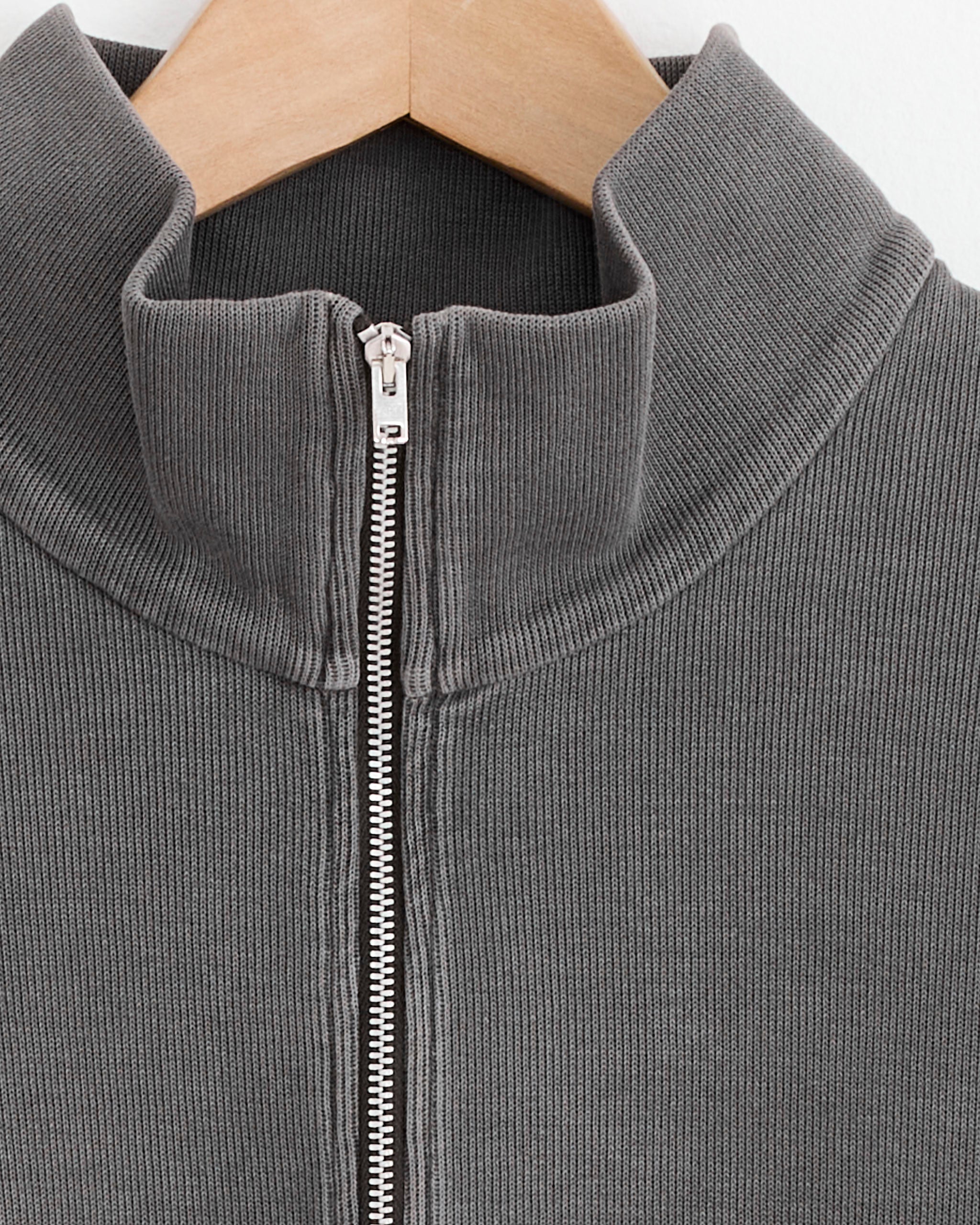 Close-up of an Our Legacy Shrunken Full Zip Sweater in Worn Graphite ribbed knit on a wooden hanger, featuring a silver zipper and mock neck collar.