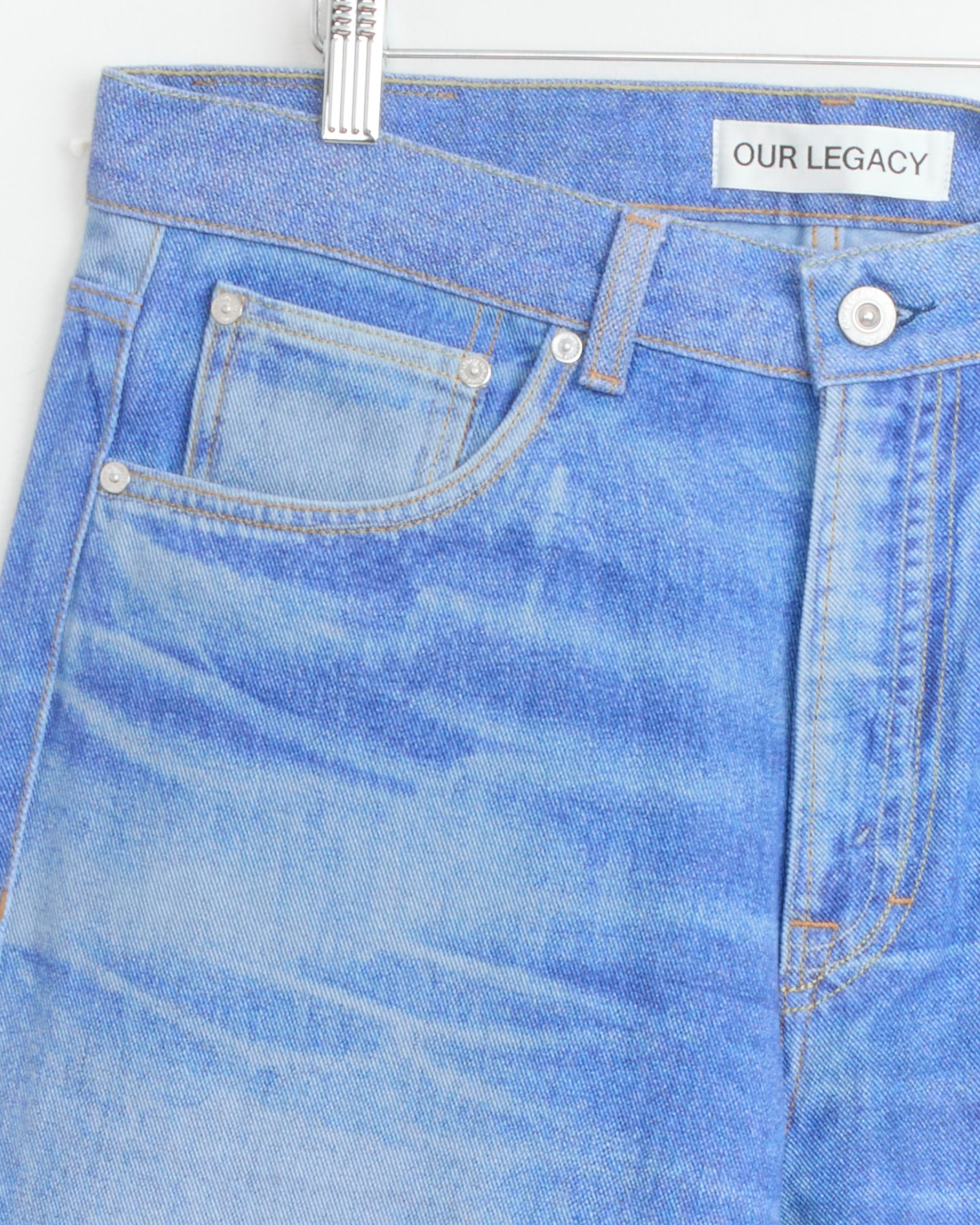 Close-up of the Third Cut Jean in Digital Blue Aurora Print by Our Legacy hanging on a white hanger, featuring classic stitching, a coin pocket, and an OUR LEGACY label inside the waistband.