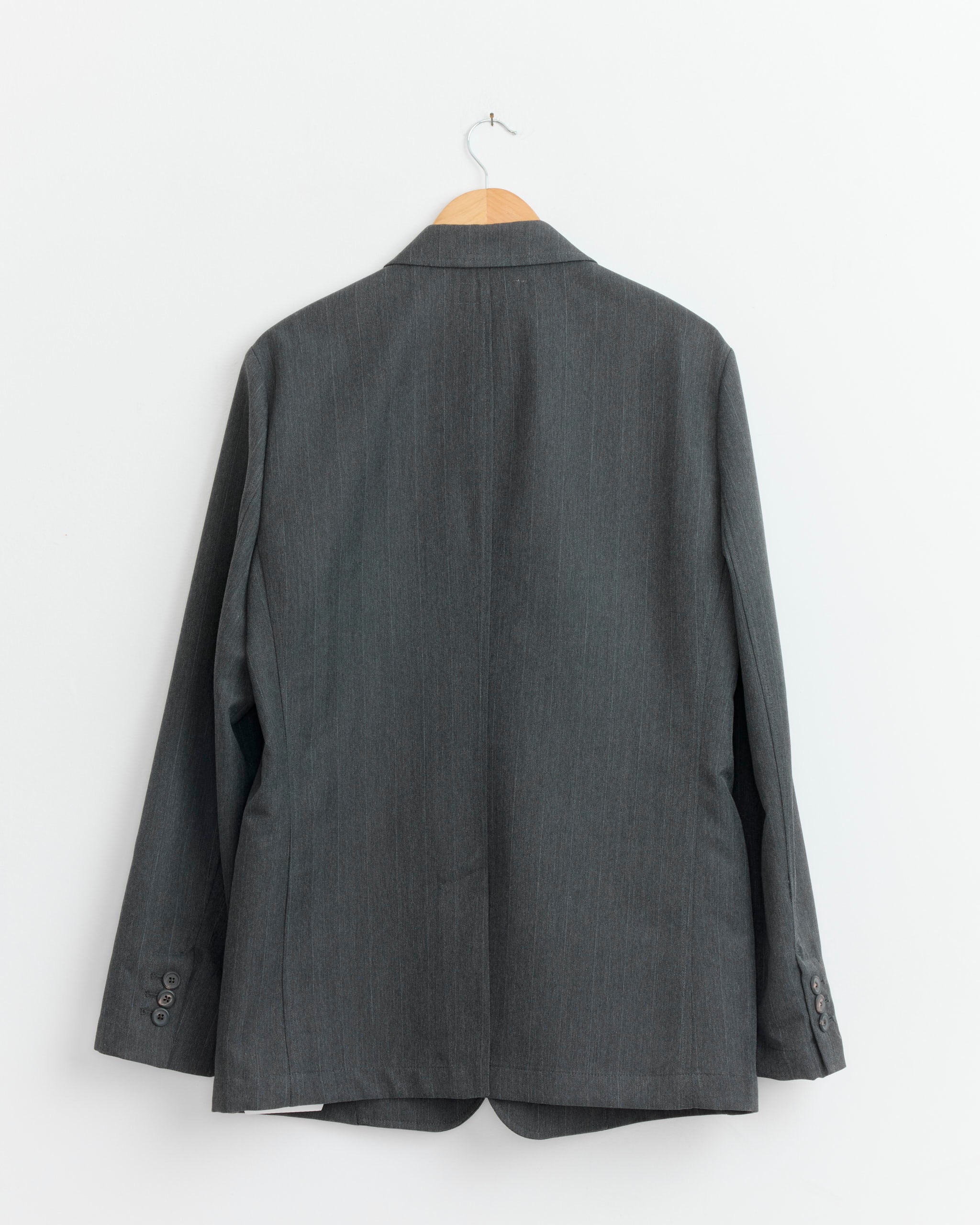 The Unconstructed Ceremony Blazer in Shark Pinstripe by Our Legacy, made from Italian suiting fabric, hangs on a wooden hanger against a white wall, displayed from the back.