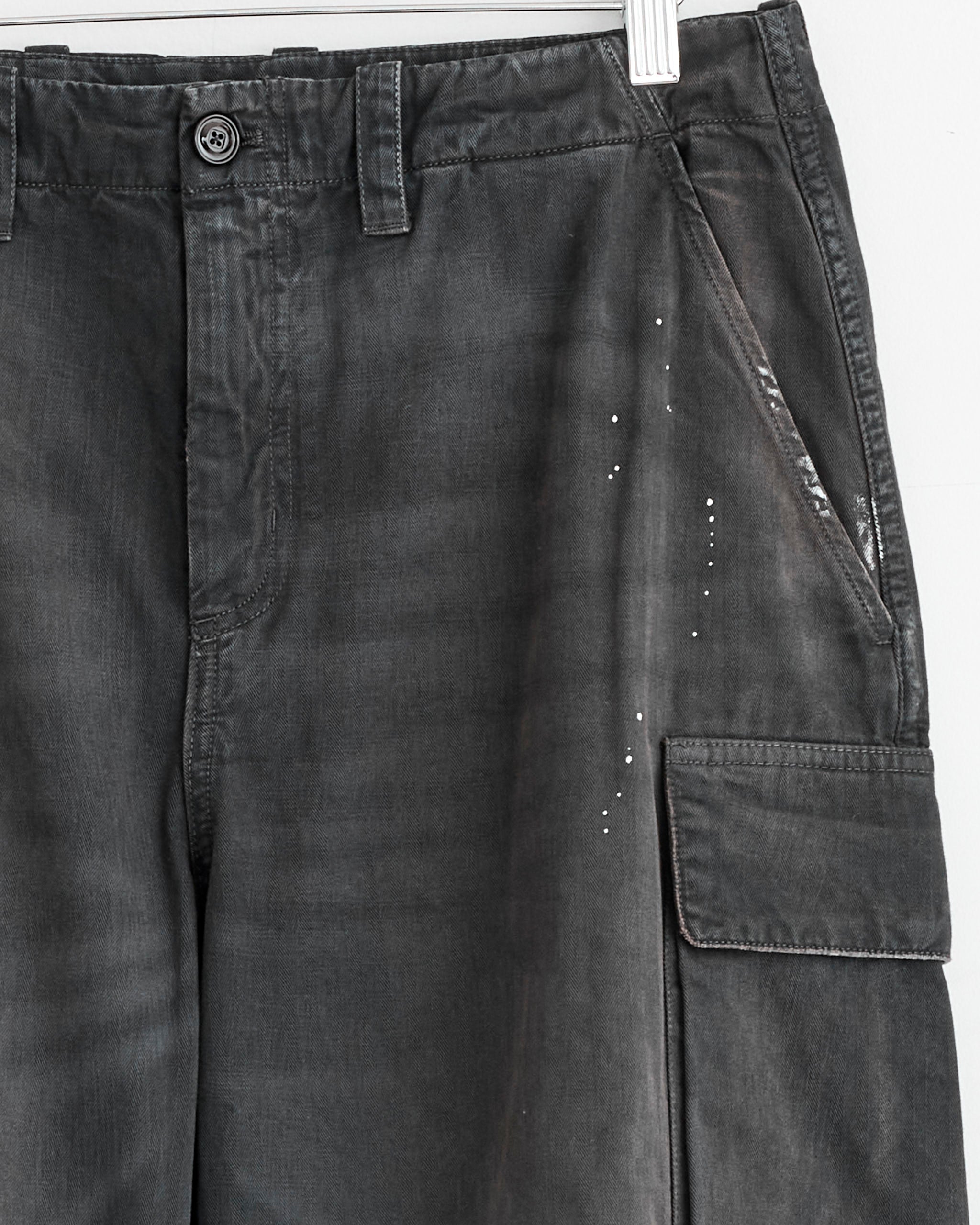Close-up of Our Legacy’s Mount Cargo Pant in Nebula Wash, Italian cotton herringbone, with side pockets, button closure, belt loops, and white paint splatters on the right thigh, hanging on a white hanger.